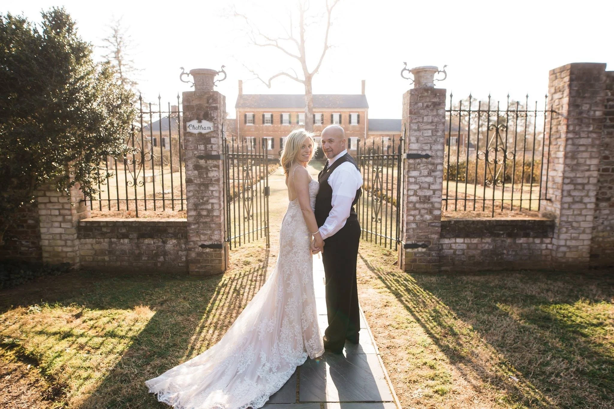 A bride and groom holding hands, standing in front of a brick gate on a sunny day, with a large historic house in the background.