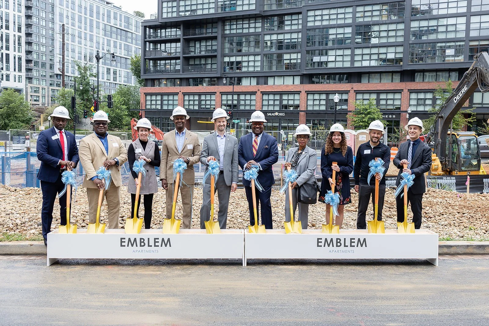 Group of men and women in business attire wearing construction helmets, holding small shovels decorated with blue ribbons, standing behind a sign that reads 'EMBLEM APARTMENTS' during a groundbreaking ceremony at a construction site in an urban area with high-rise buildings in the background.