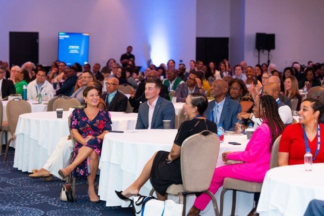 Audience seated at round tables during a conference or seminar in a large, well-lit meeting room.