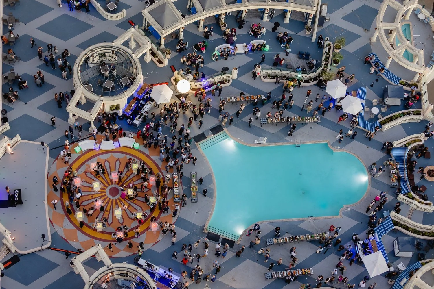 An aerial view of a busy outdoor shopping mall or entertainment area with a large swimming pool, a colorful circular seating area, and numerous people walking and sitting. The space includes small gathering spots, cafes with umbrellas, and retail sta