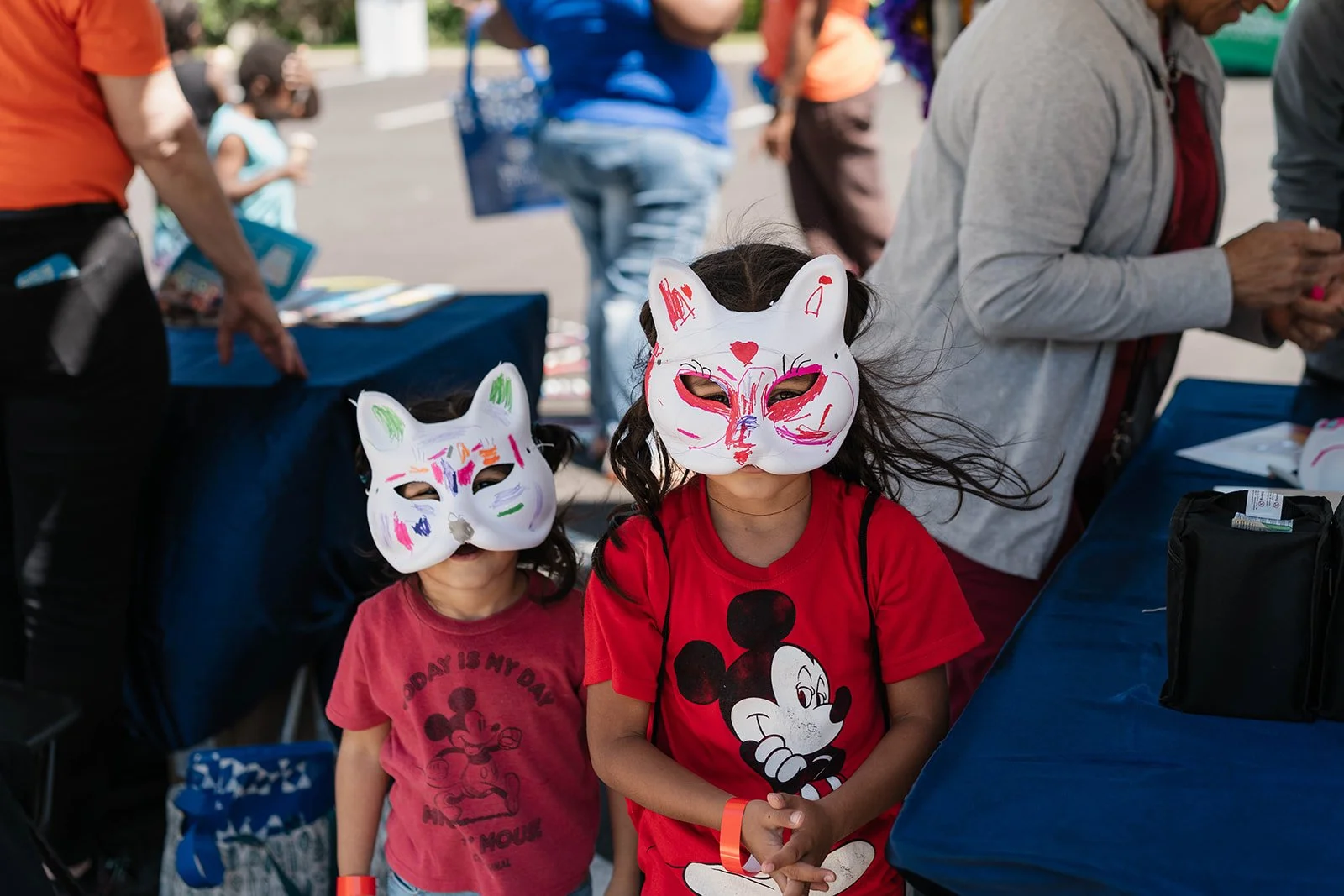 Two children wearing white animal masks with colorful drawings stand at an outdoor event. One child wears a red Mickey Mouse shirt, and the other wears a pink shirt with Mickey Mouse on it. People are seen in the background around a table with blue t