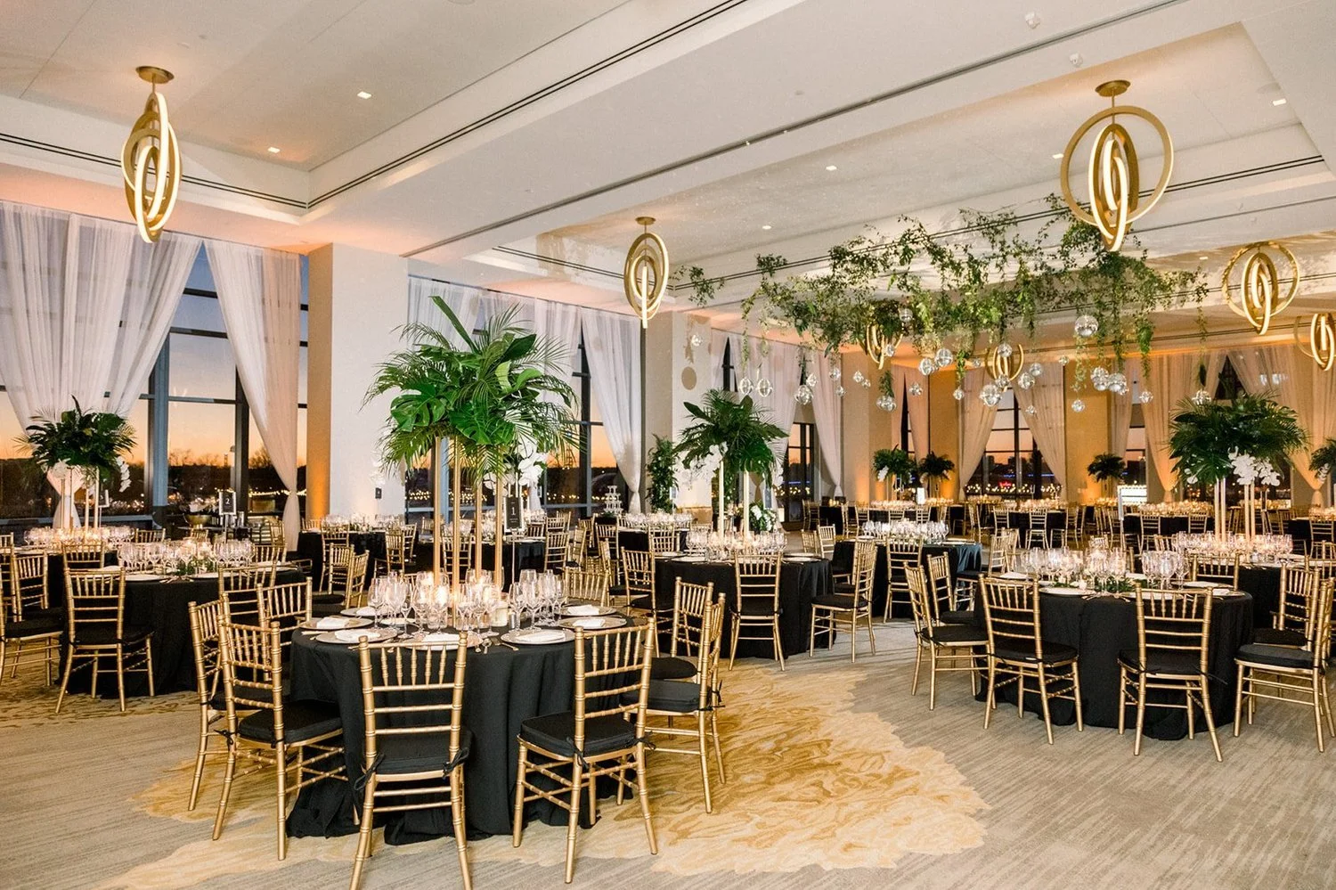Elegant banquet hall decorated with gold chairs, black tablecloths, large green plants, and hanging gold and glass light fixtures, with large windows showing a sunset view.