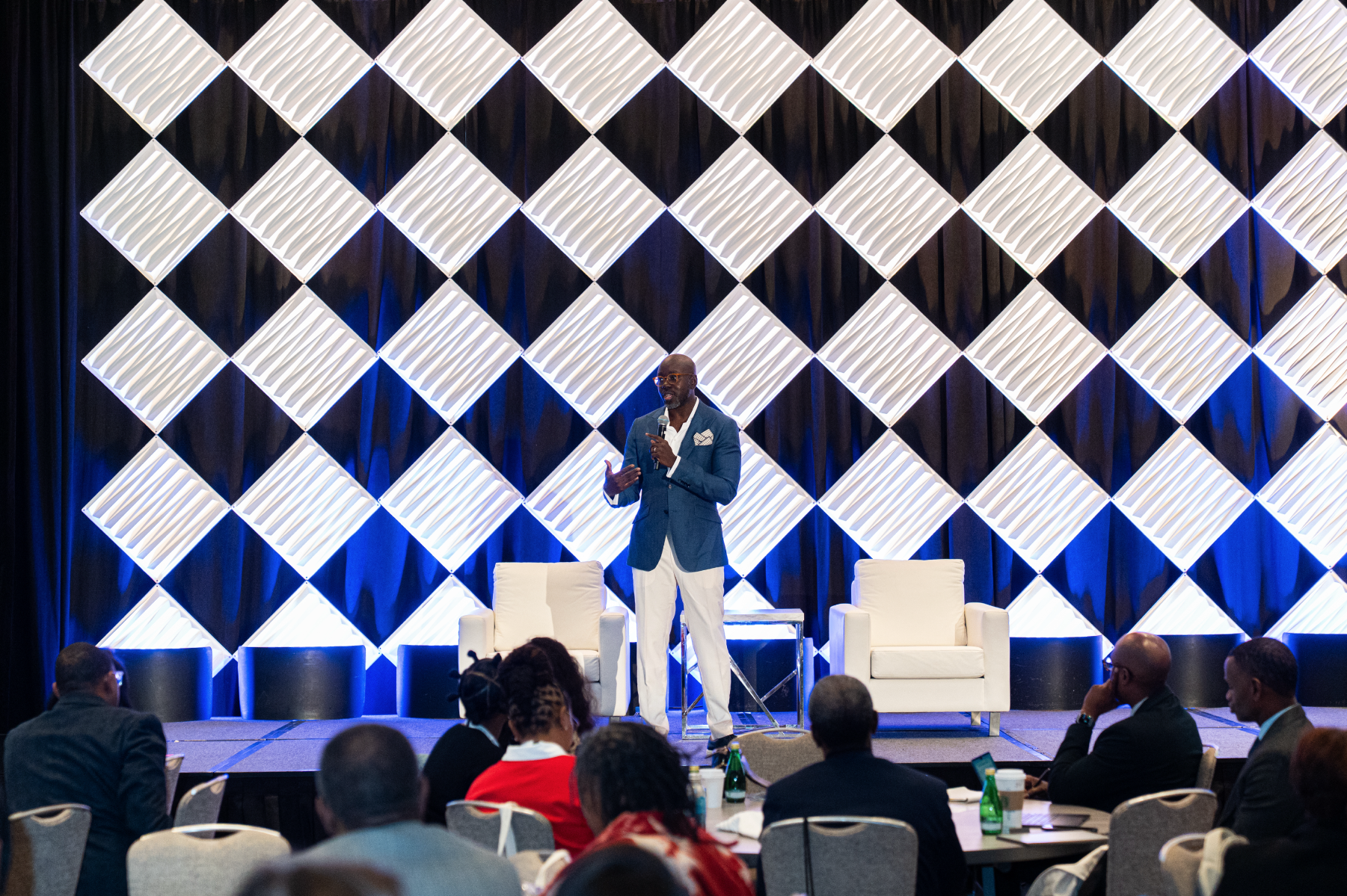 A man in a blue blazer and white pants speaking into a microphone on a stage with a geometric lit backdrop, while seated audience watches.