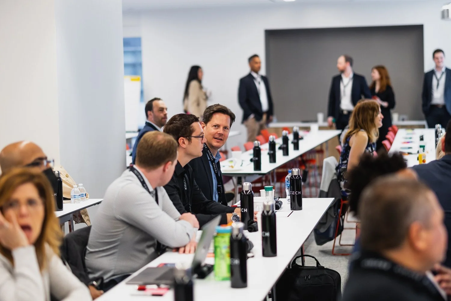 People attending a business workshop or conference in a modern conference room, sitting around long white tables with water bottles, laptops, and notebooks, some standing and engaging in conversation at the back.
