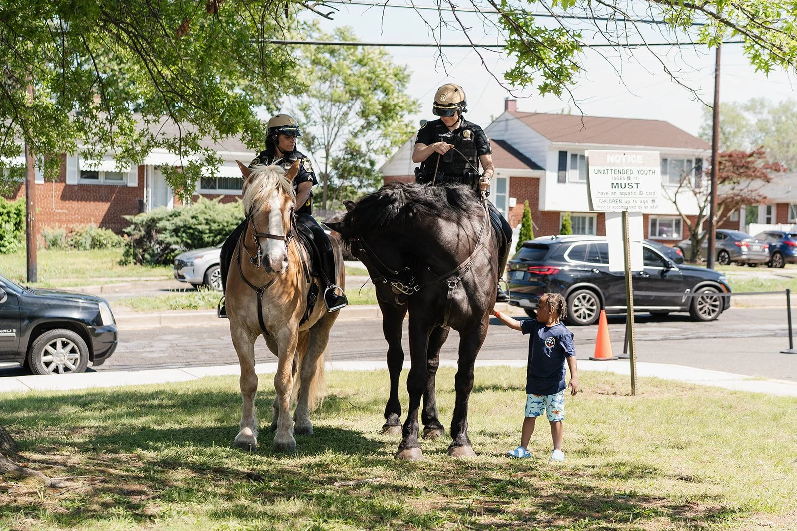 Two police officers on horseback interact with a young boy holding a small animal by a leash in a park. The scene is shaded by trees, with cars and houses visible in the background.