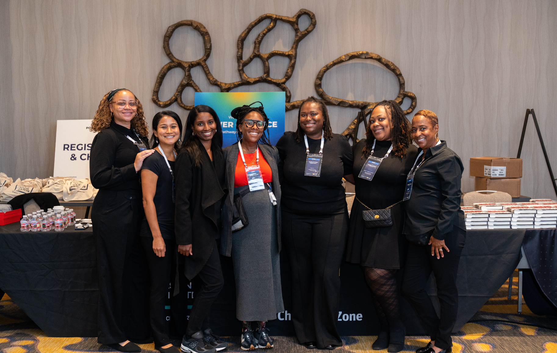 Group of seven women posing together at a conference or event, standing in front of a registration table with conference materials, in a conference hall or hotel ballroom.