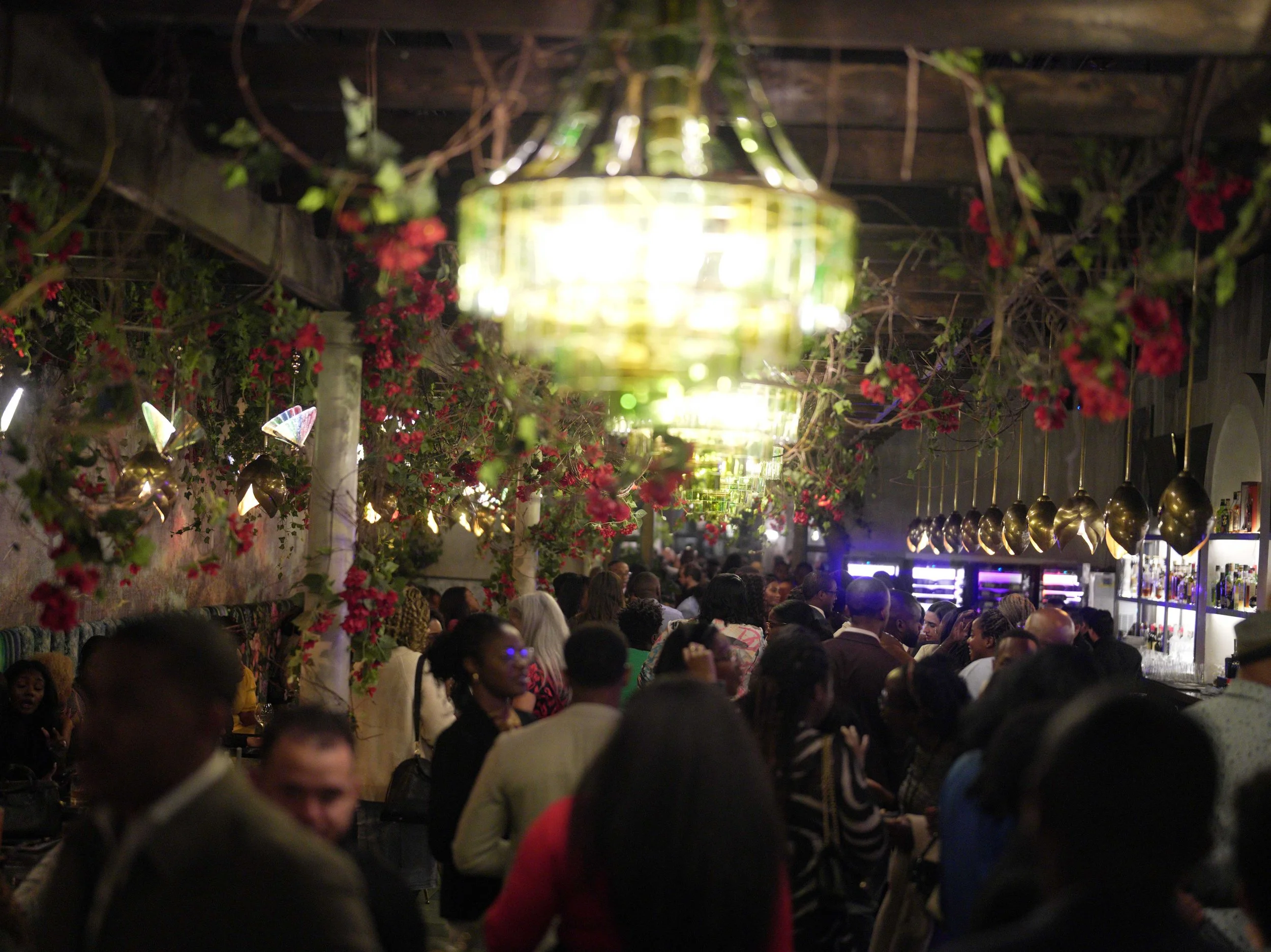 A crowded indoor bar or restaurant with people sitting and socializing, decorated with hanging golden pendant lights and lush green plants with red flowers.
