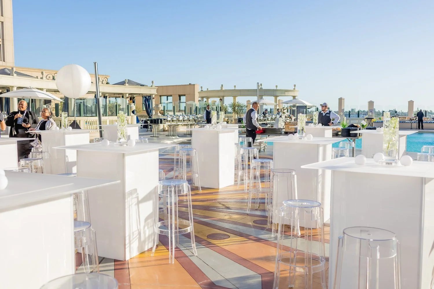 An outdoor rooftop party setup with white tables and clear acrylic stools, elegant floral centerpieces, and staff preparing food, overlooking a cityscape in the background.