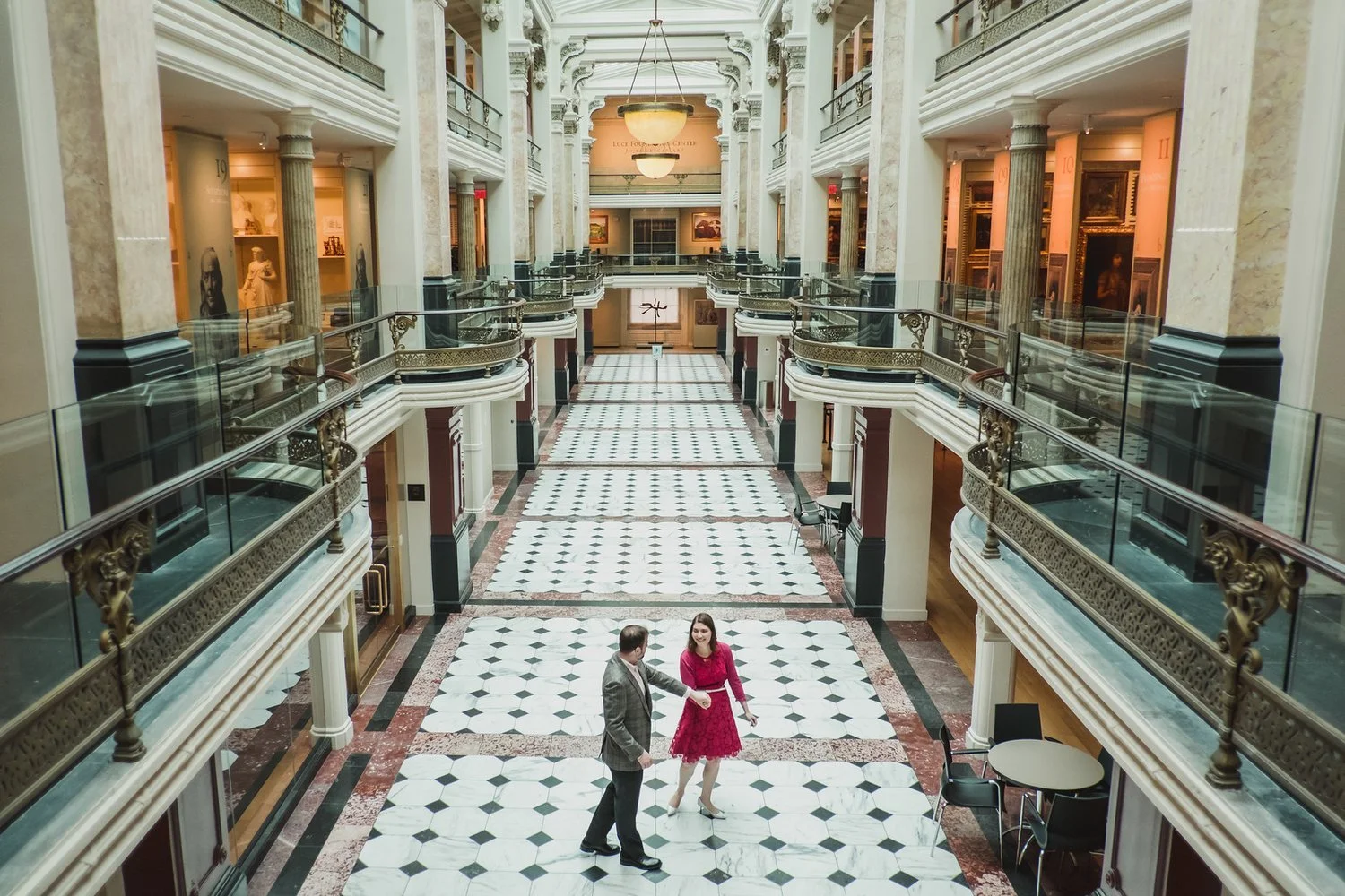 Interior view of a grand, multi-story building with a marble floor, ornate columns, and wall art. Two people are dancing in the center of the lobby.