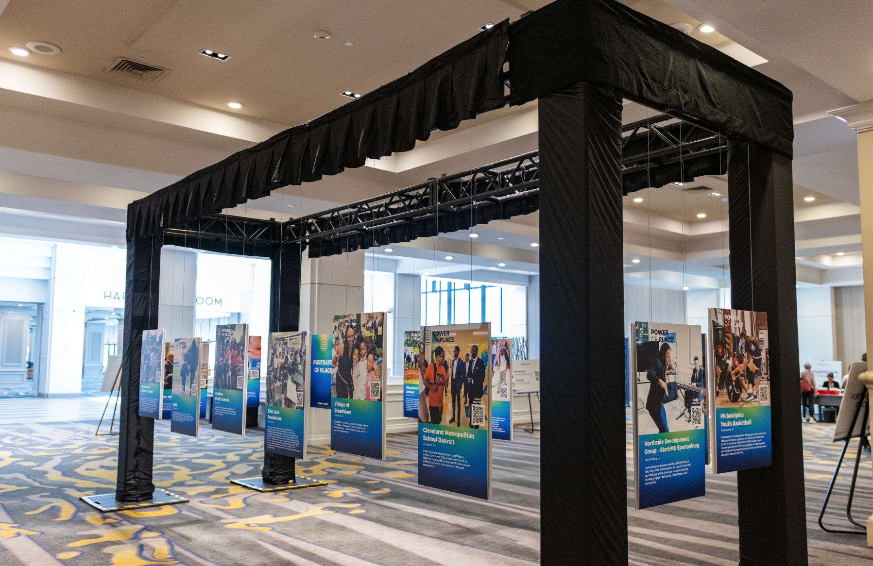 Indoor display with posters on stands under a black canopy in a spacious building lobby. The posters feature diverse groups of people and various community and school programs.