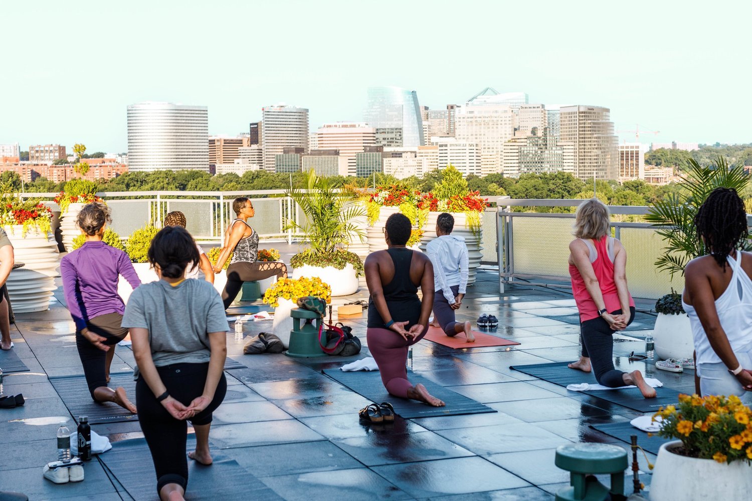 People participating in an outdoor yoga class on a rooftop with a city skyline in the background, surrounded by potted plants and flowers.