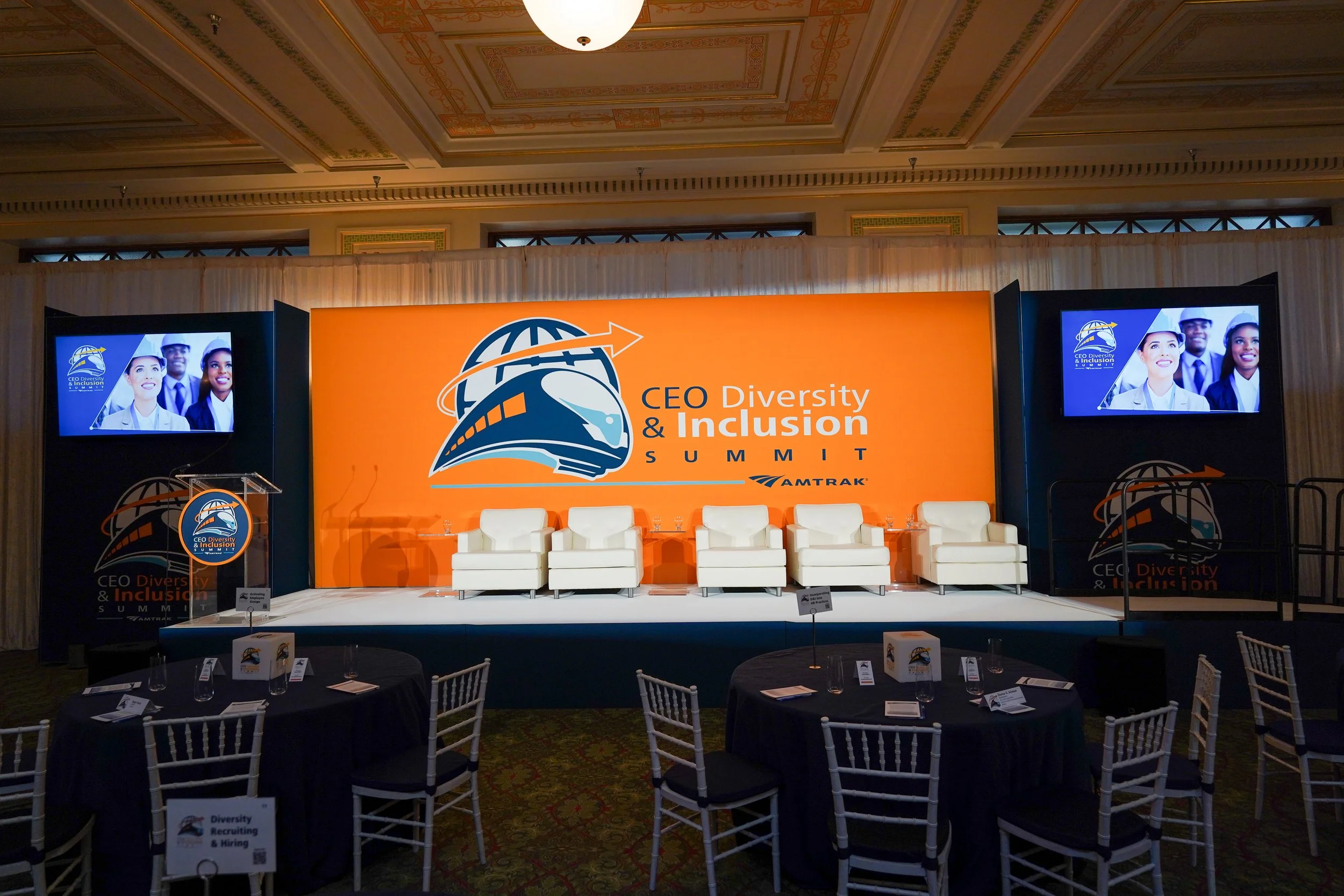 A stage set up for the CEO Diversity & Inclusion Summit with white chairs, a large orange banner displaying the event logo, and two screens showing the event logo and images of diverse professionals in business attire. The ceiling has ornate detailin