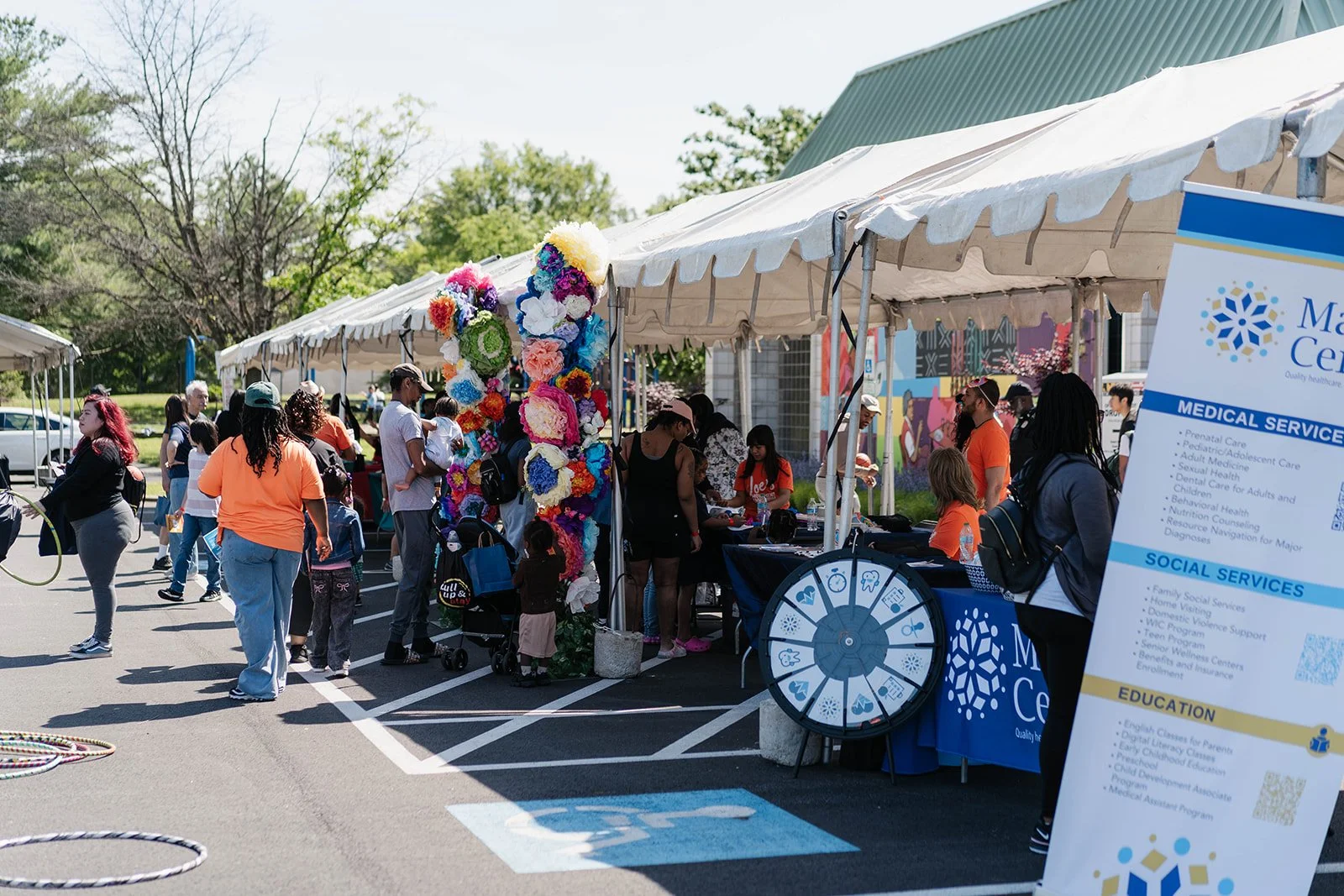 People gathered at outdoor community event with vendor booths, colorful decorations, and informational displays, on a bright day with trees and a mural in the background.