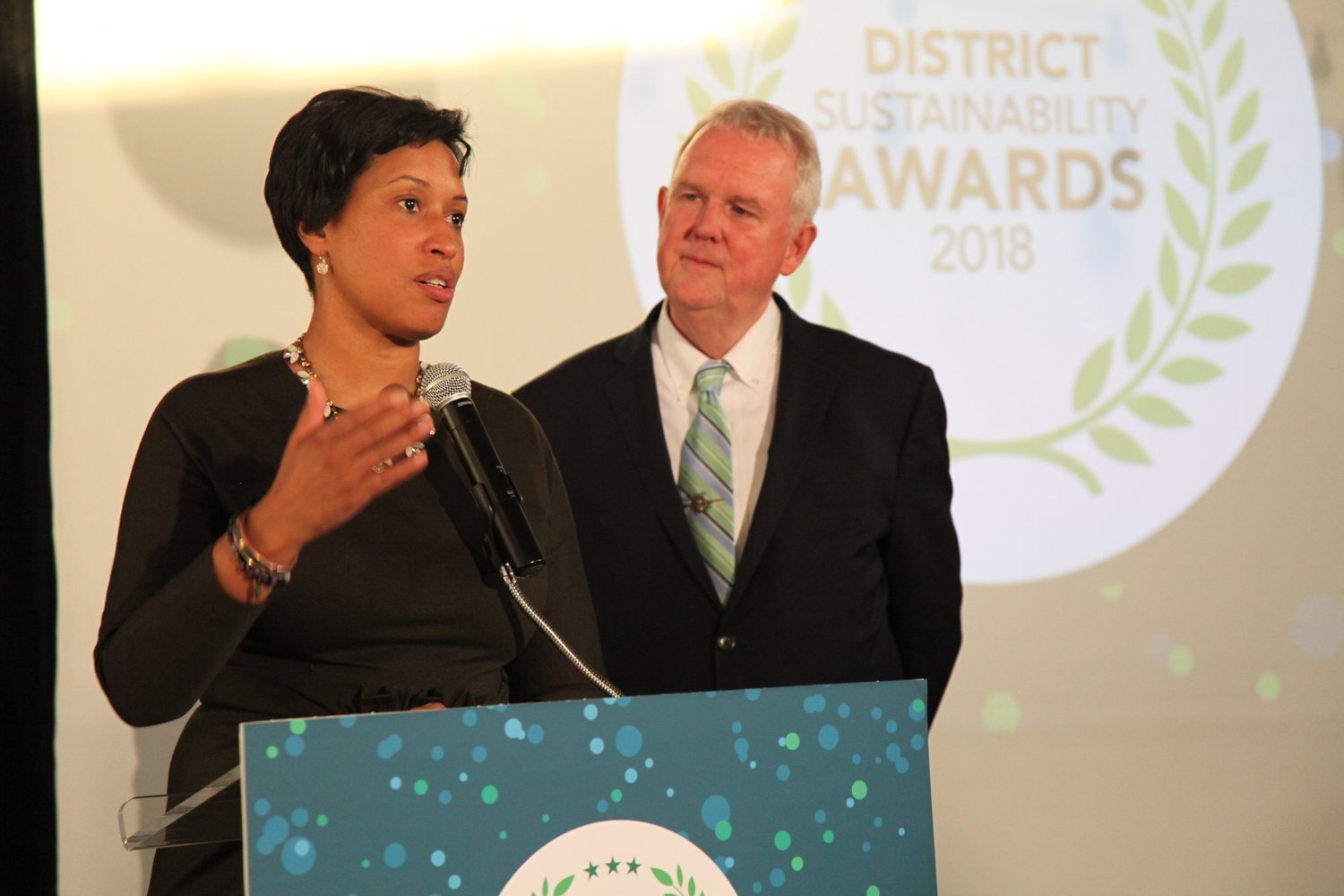 A woman speaking at a podium during an award ceremony, with a man standing behind her, and a round sign in the background that reads 'District Sustainability Awards 2018'.