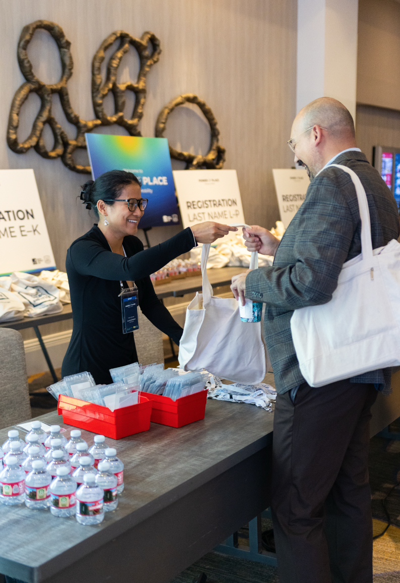 A woman at a registration table handing a badge to a man holding a tote bag, with water bottles and supplies on the table and registration signs in the background.
