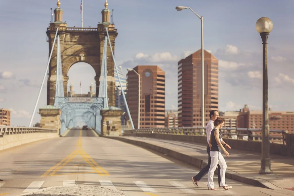 A couple walking across a bridge in an urban area, with tall city buildings in the background, under a partly cloudy sky.