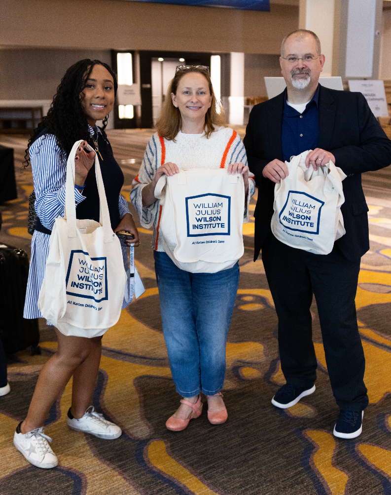 Three people standing inside an airport or conference center, holding tote bags with the William Julius Wilson Institute logo. The woman on the left has curly black hair, wearing a striped blue and white dress, and white sneakers. The woman in the mi
