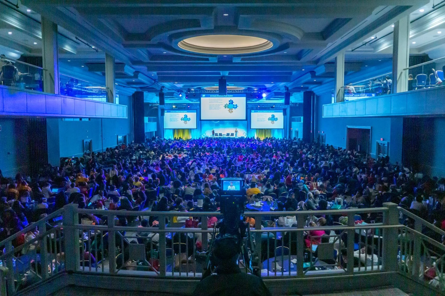 Large conference hall filled with a seated audience attending a presentation on stage with multiple screens showing a slide titled 'S-Phony Oklahoma State System'.