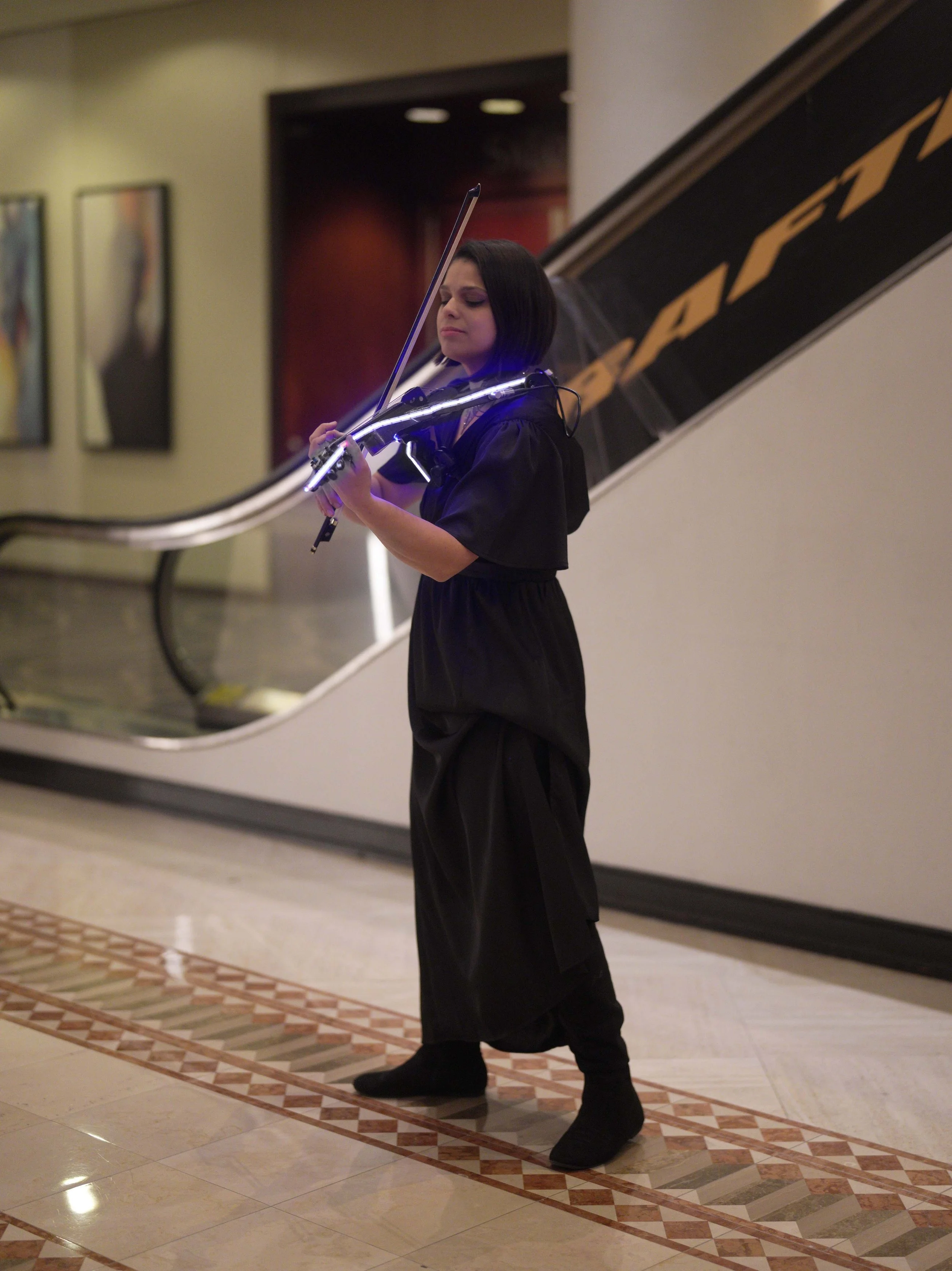 A woman with short black hair, wearing a black outfit and boots, is standing indoors near a staircase, playing a violin with neon lights on the instrument.