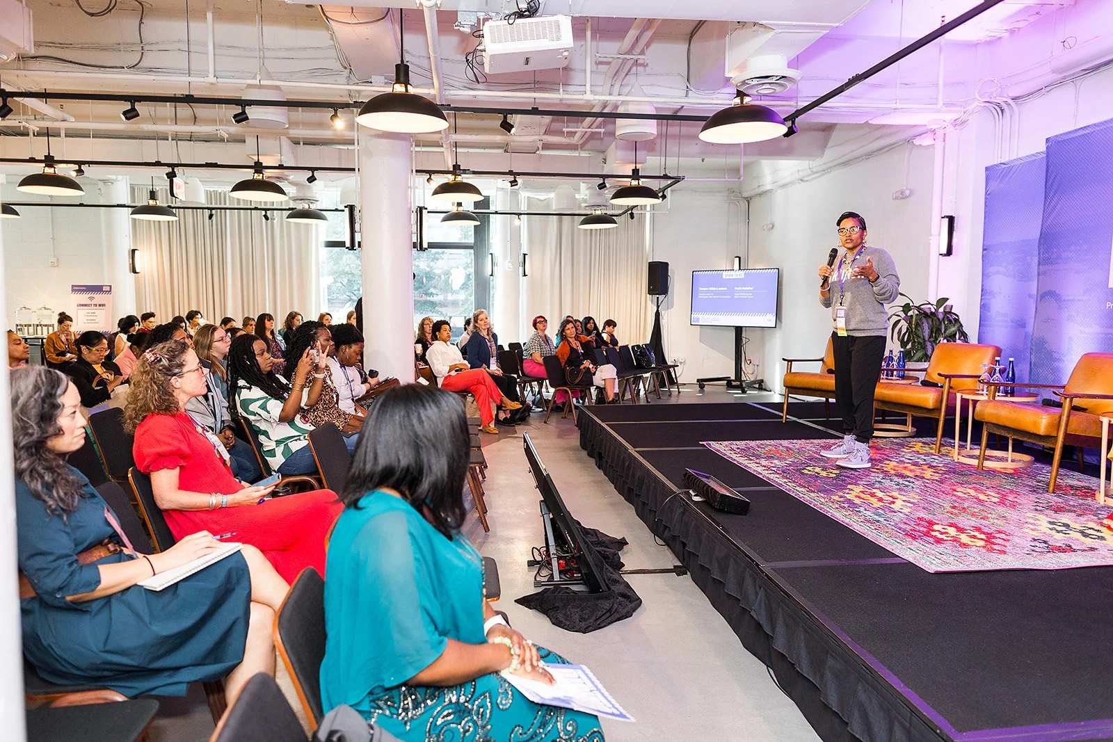 A woman is giving a presentation on a stage in front of an audience in a conference room. The audience is seated and attentively listening.