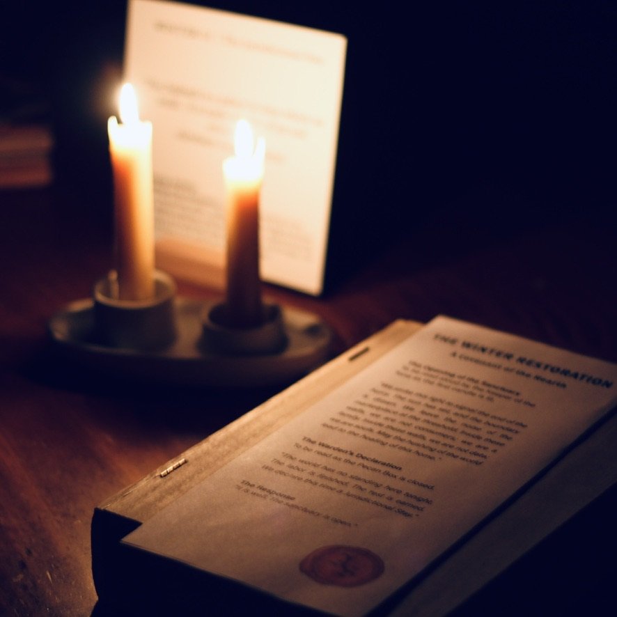 A Sabbath table set with two beeswax taper candles, a seasonal liturgy atop a wooden box and weekly rhythm card placed in a wooden block holder.  The scene is warm and peaceful.