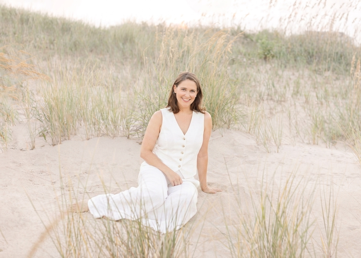 A woman in a white outfit sitting on sandy beach with tall grasses around her and distant dunes in the background, smiling at the camera.