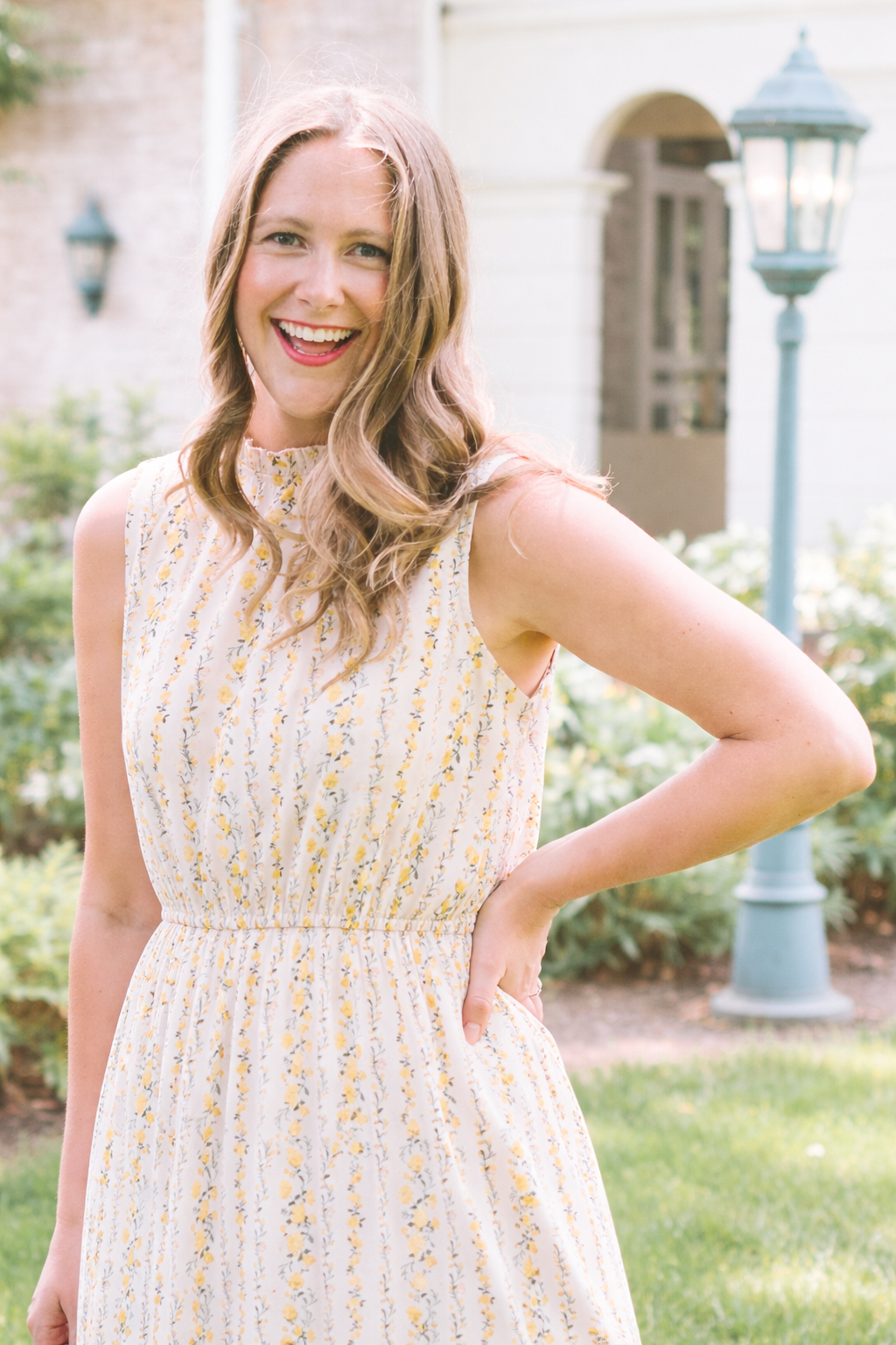 A smiling woman in a sleeveless, floral dress standing outdoors near a lamp post.