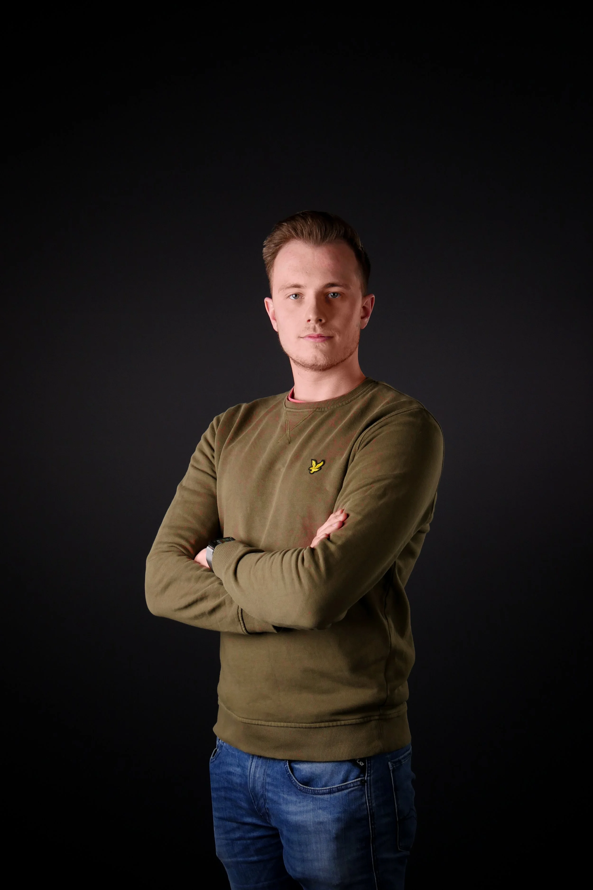 Portret van een jonge man met een groene trui en blauwe jeans, met gekruiste armen, staand voor een zwarte achtergrond.