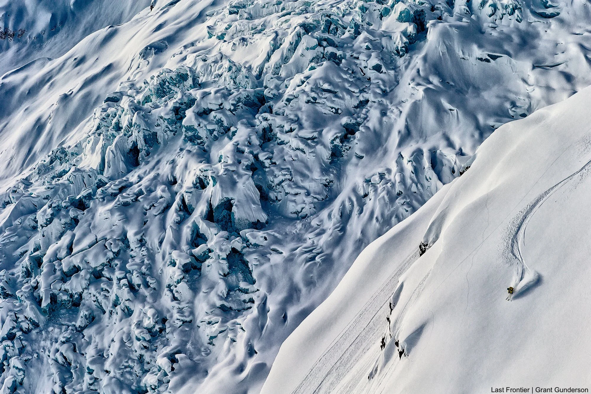 Skier making turns on a broad alpine shoulder with a glacier and rugged Coast Mountain peaks in the background.