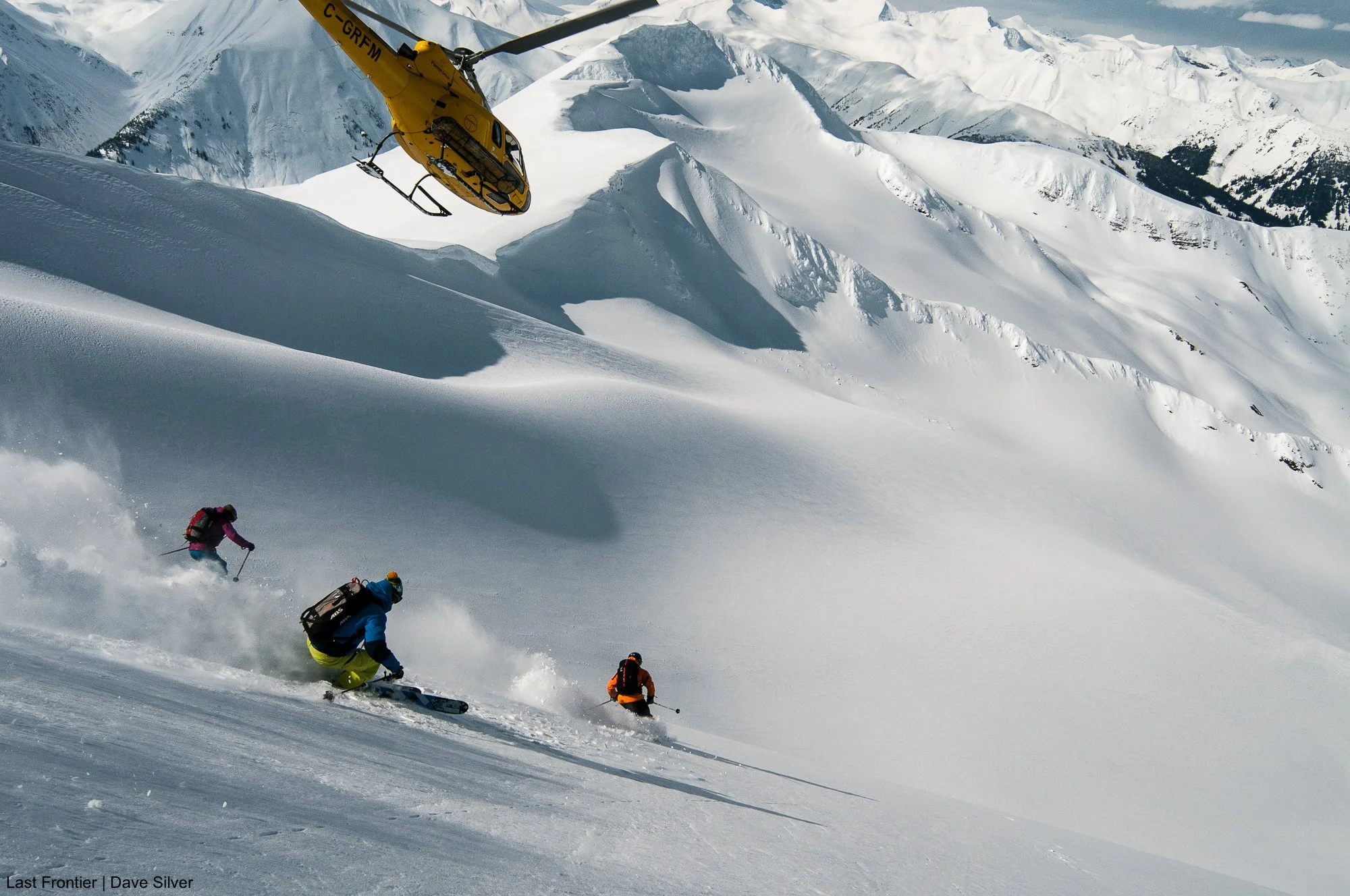 Heli-skiers descending alpine terrain at Bell 2 Lodge with a Last Frontier helicopter overhead.