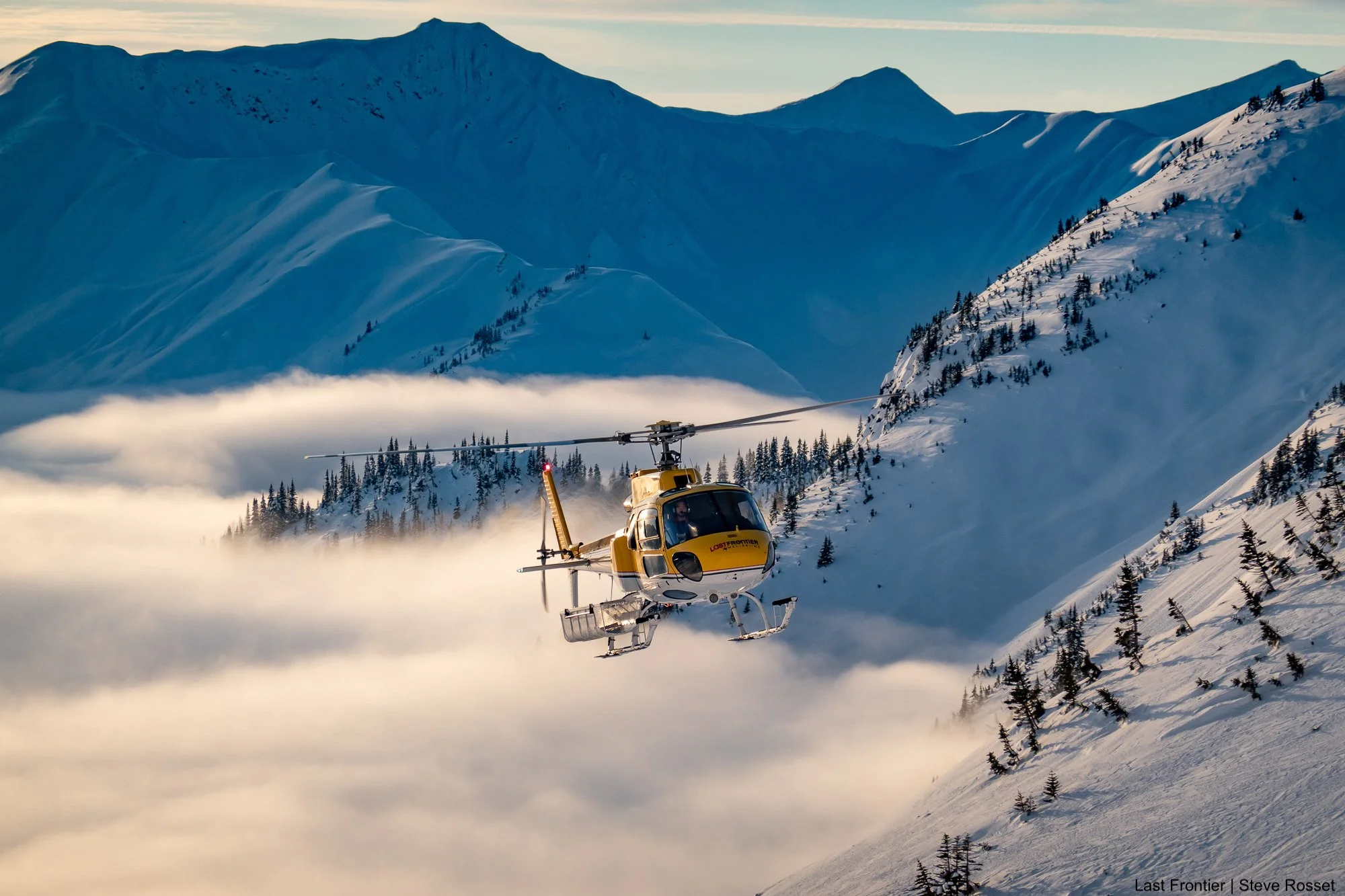 Last Frontier Bell 2 helicopter flying above the mountains in golden evening light.
