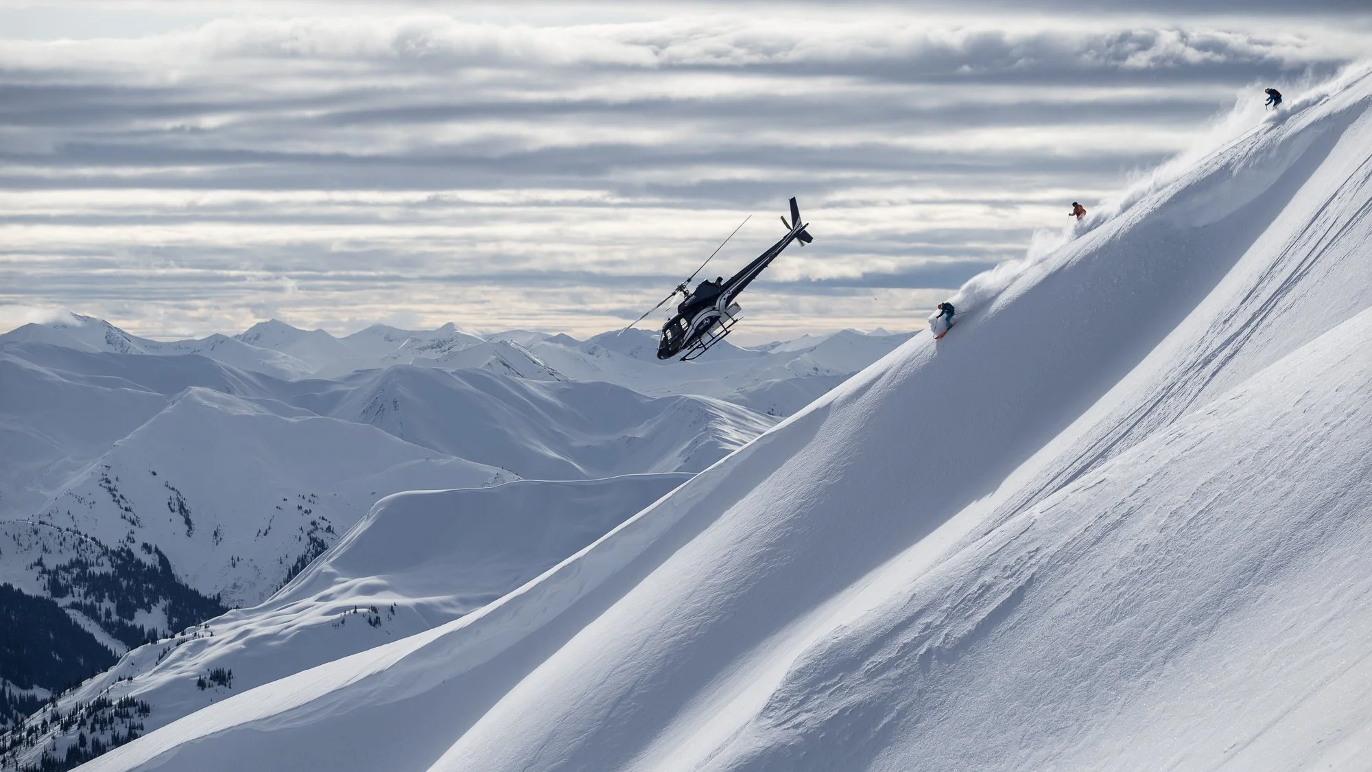 heli skier descends from alpine peak with helicopter flying over head at Last frontier heliskiing bell 2 lodge