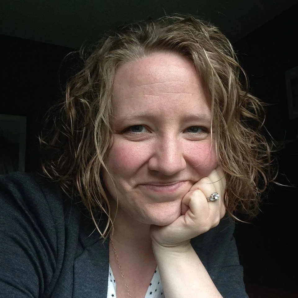 A woman with shoulder-length curly hair, smiling with her head resting on her hand, wearing a dark top and a ring on her ring finger.