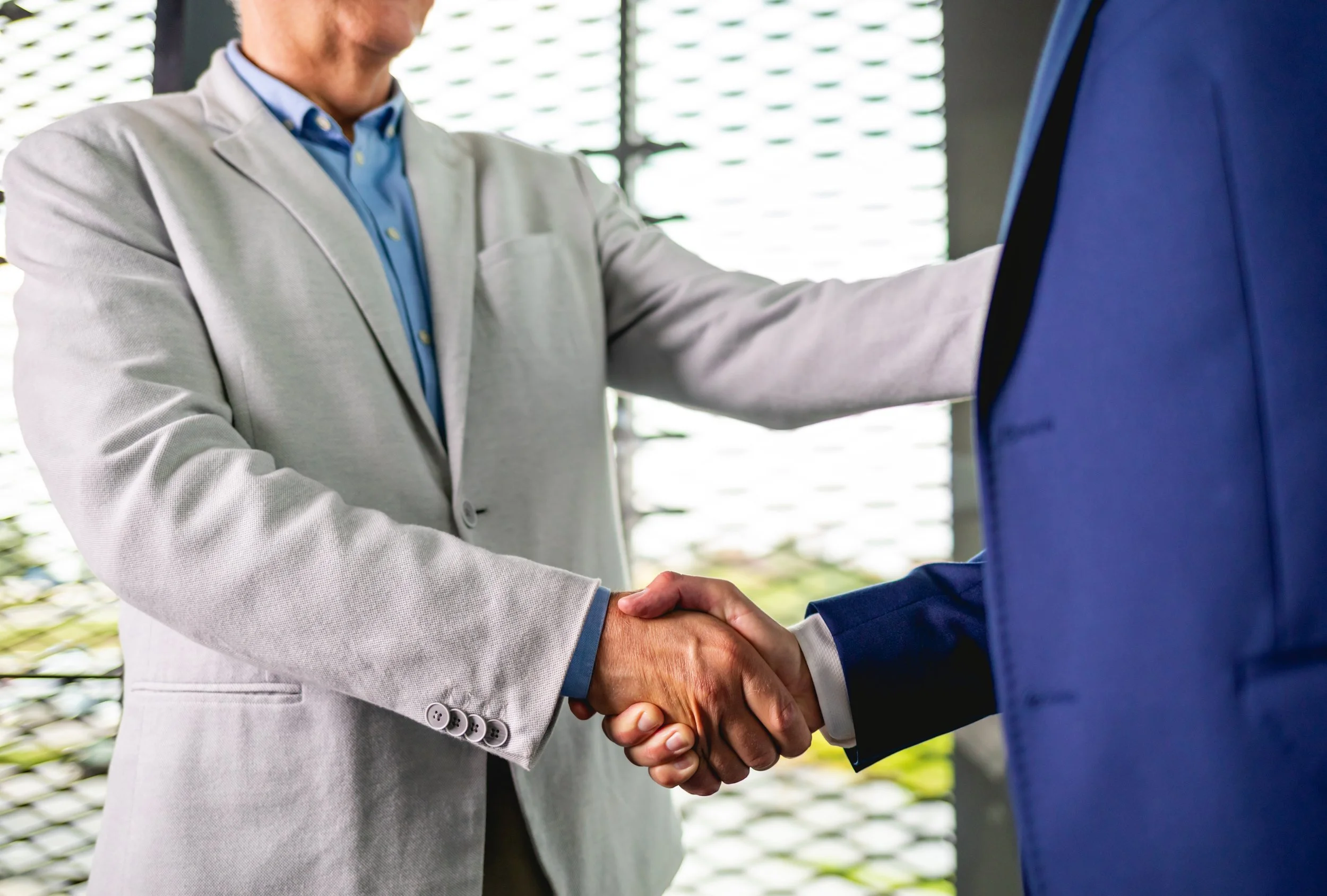 Two individuals in business suits shaking hands in front of a window with blinds.