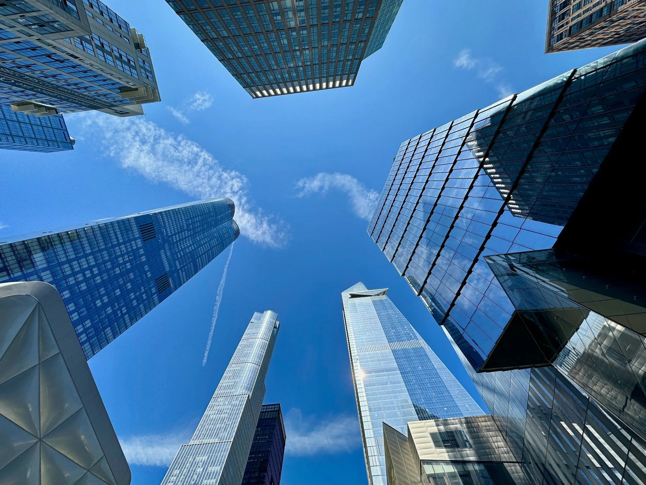 Looking up at tall modern skyscrapers with glass facades in a city under a blue sky with scattered clouds.