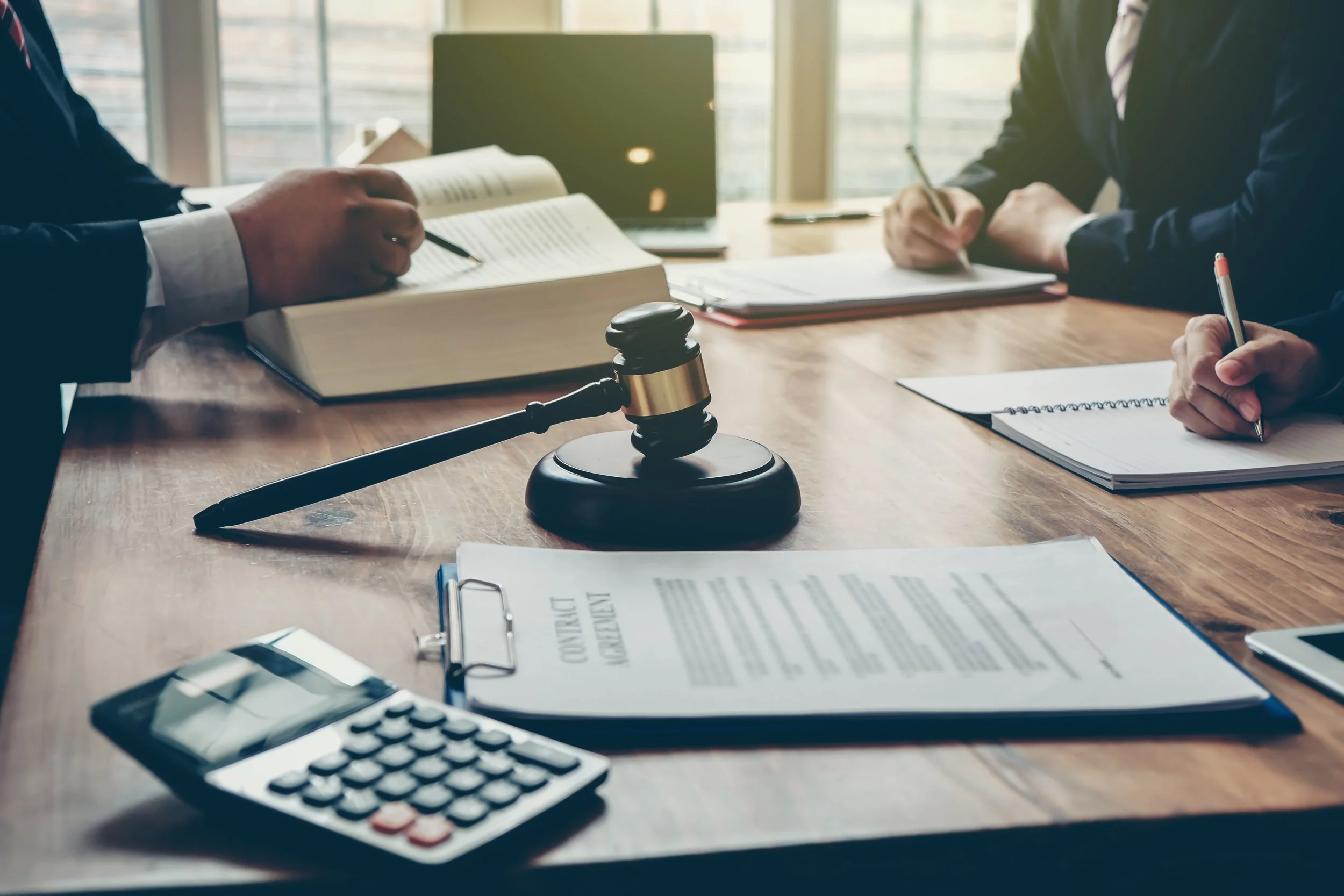 A gavel sits on a wooden table surrounded by legal documents, a calculator, and notebooks, with two people in suits taking notes during a legal or business meeting.