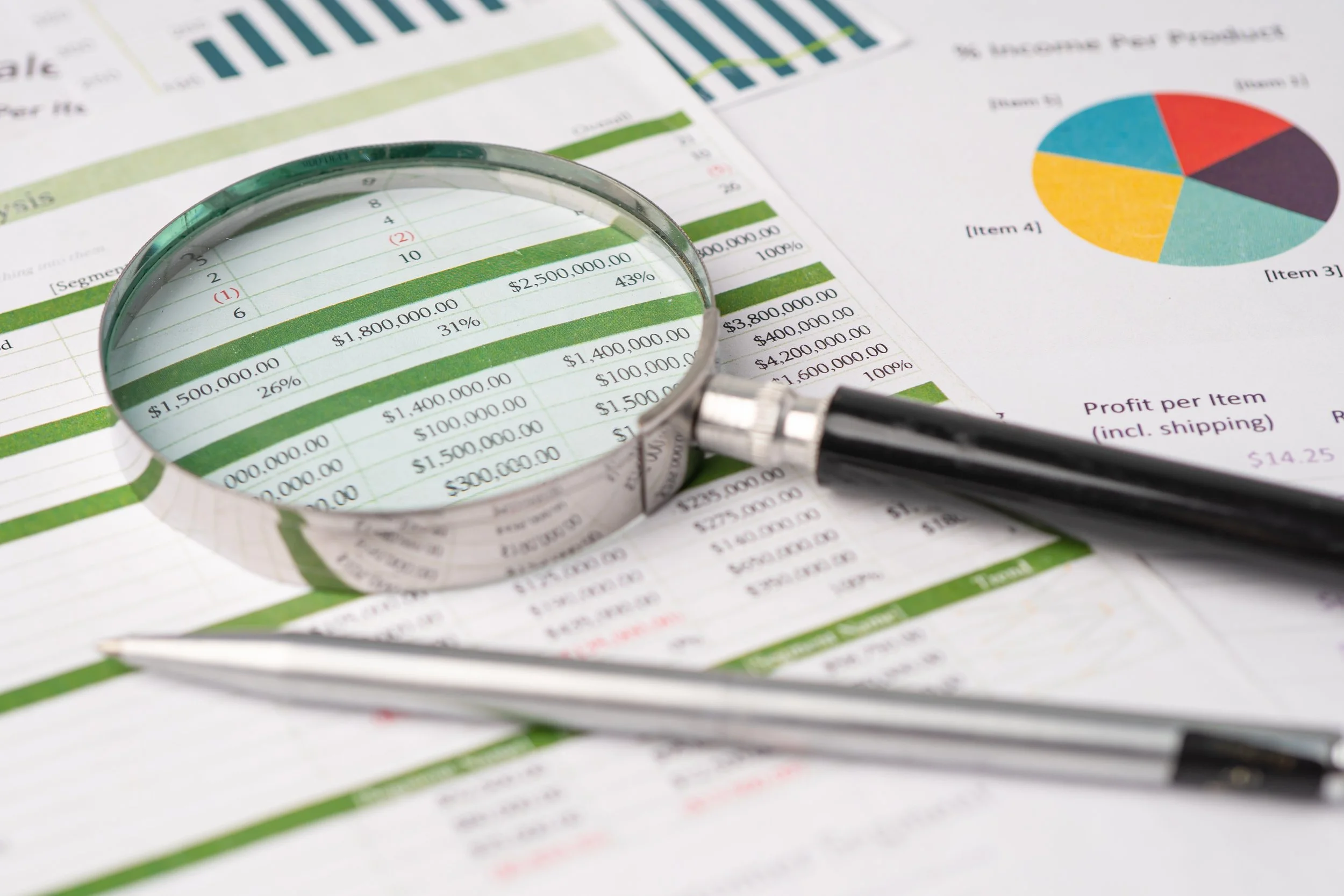 Close-up of financial reports with a magnifying glass focusing on dollar amounts and percentages, a pen, and colorful pie charts.