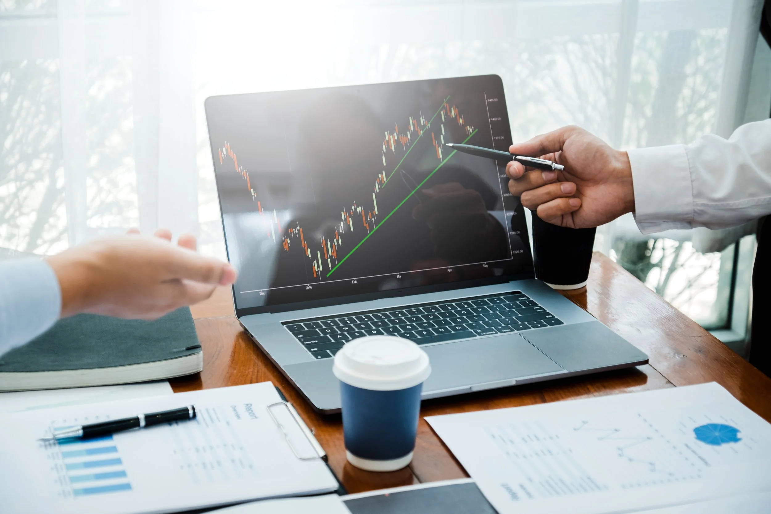 Hands pointing at a financial chart on a laptop screen in an office setting, with documents and a coffee cup nearby.