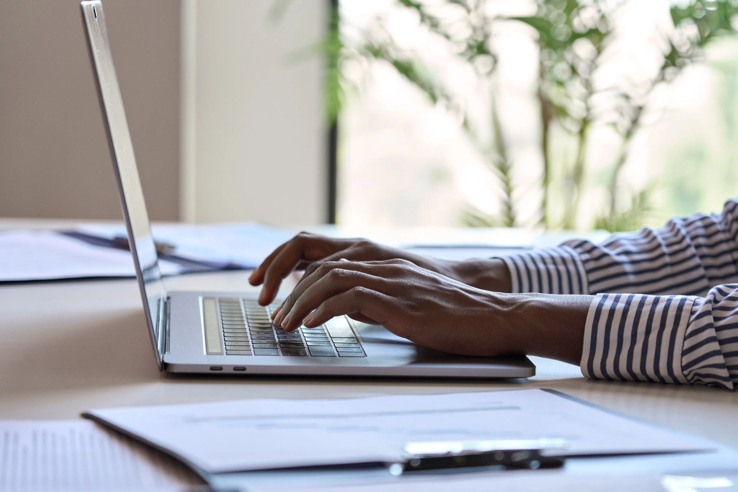 Person typing on a laptop keyboard at a desk with papers and a pen, with a blurred green plant in the background.