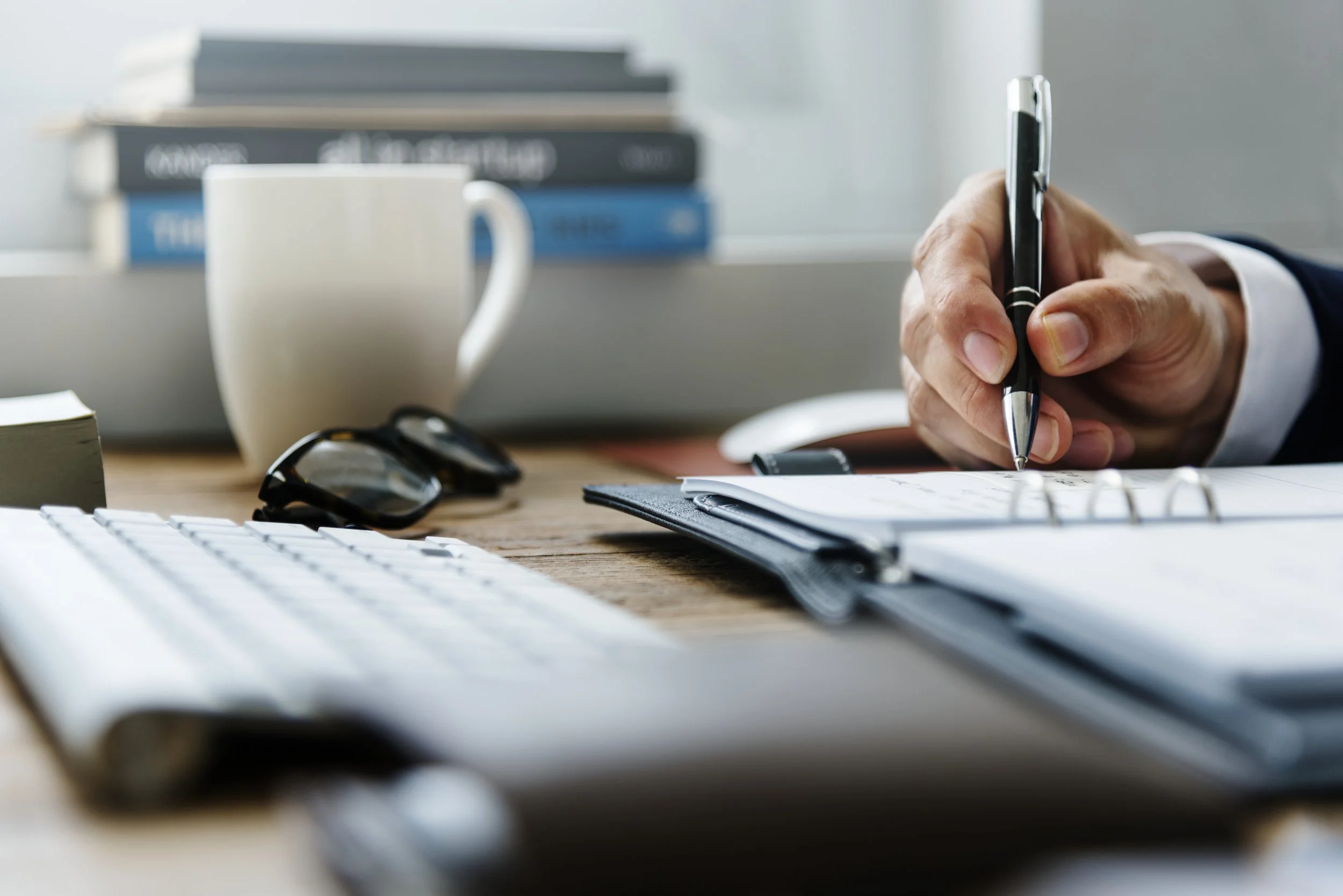 A person writing on a notepad at a cluttered wooden desk, with a computer keyboard, sunglasses, a mug, and a stack of books in the background.