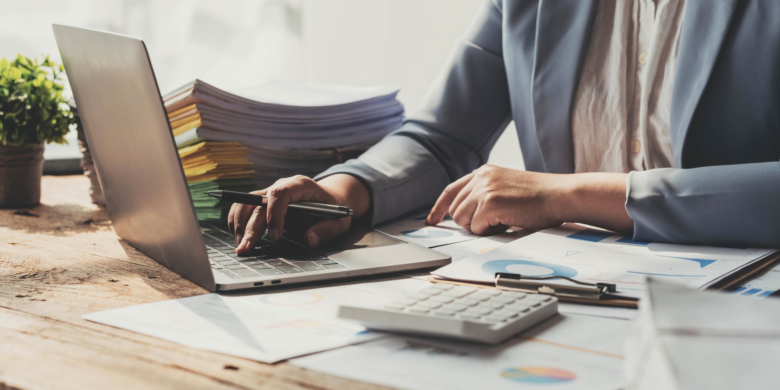 Person working at a cluttered desk with papers, a laptop, a calculator, and a pen.