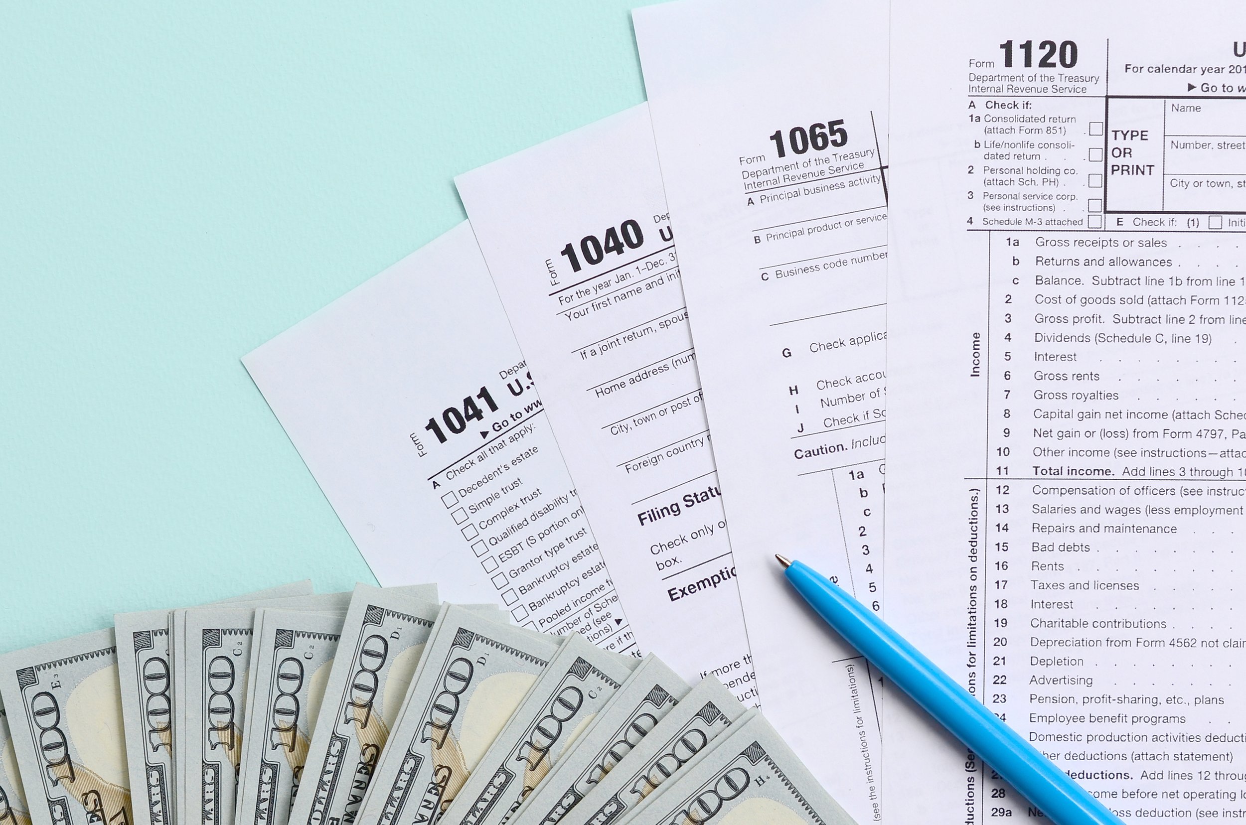 Several hundred-dollar bills fanned out next to tax forms and a blue pen on a light blue surface.