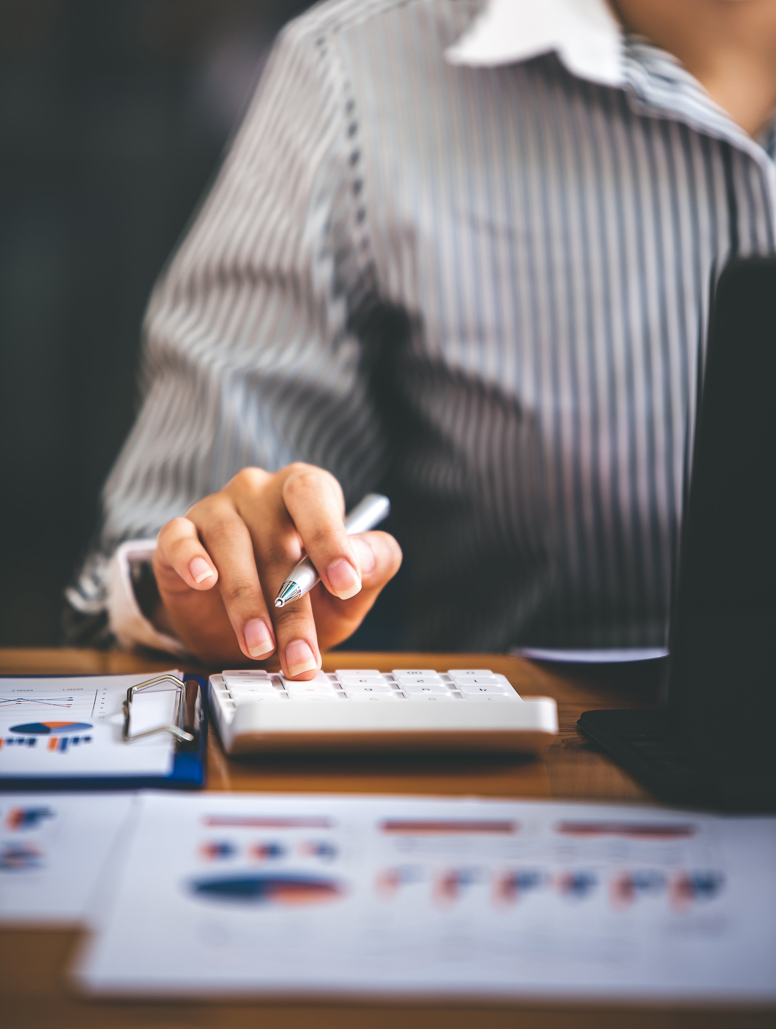 Person in a striped shirt using a calculator and a pen on a desk with charts and documents.