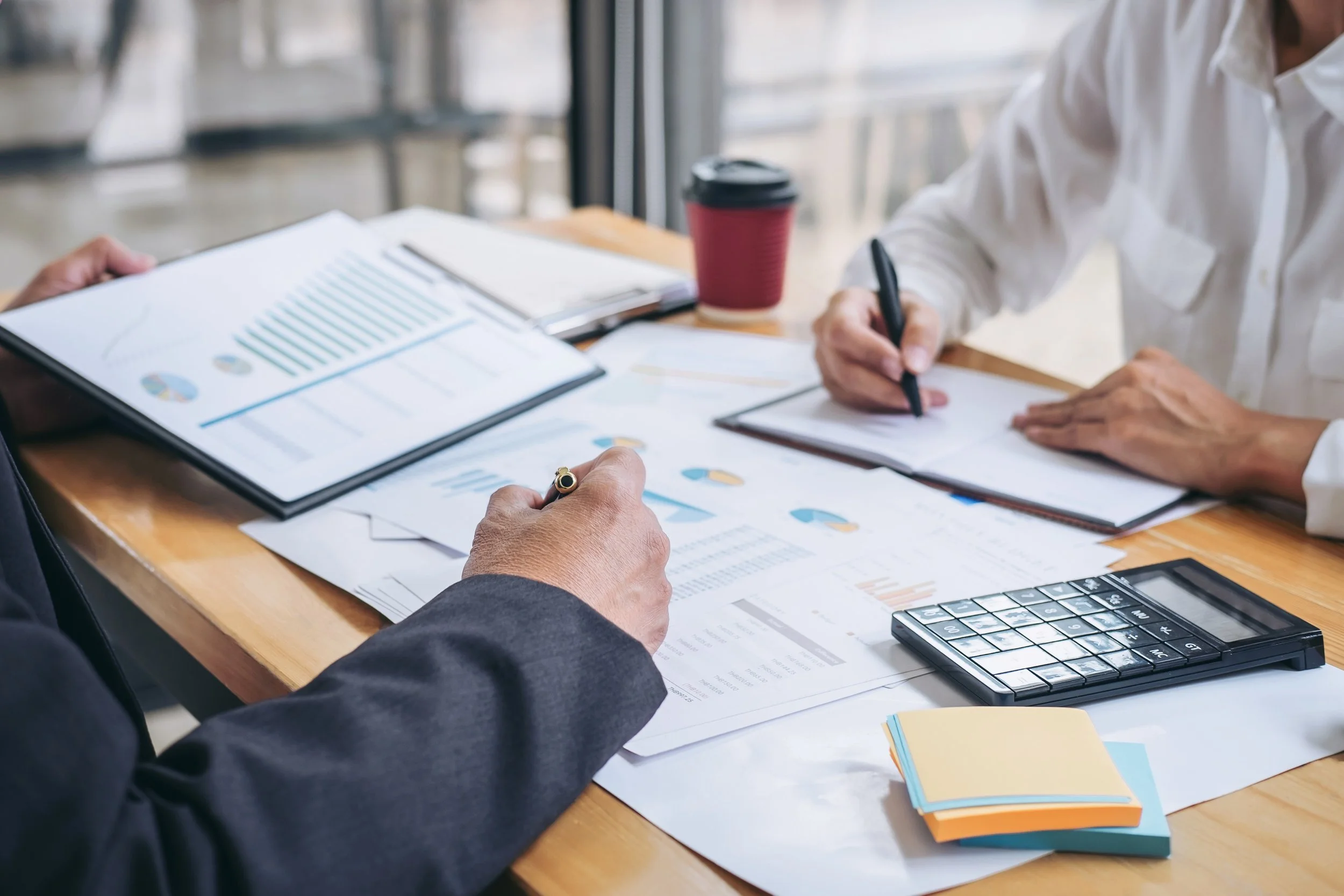 Two people sitting at a wooden table reviewing documents, charts, and graphs with a calculator, notebooks, and sticky notes.