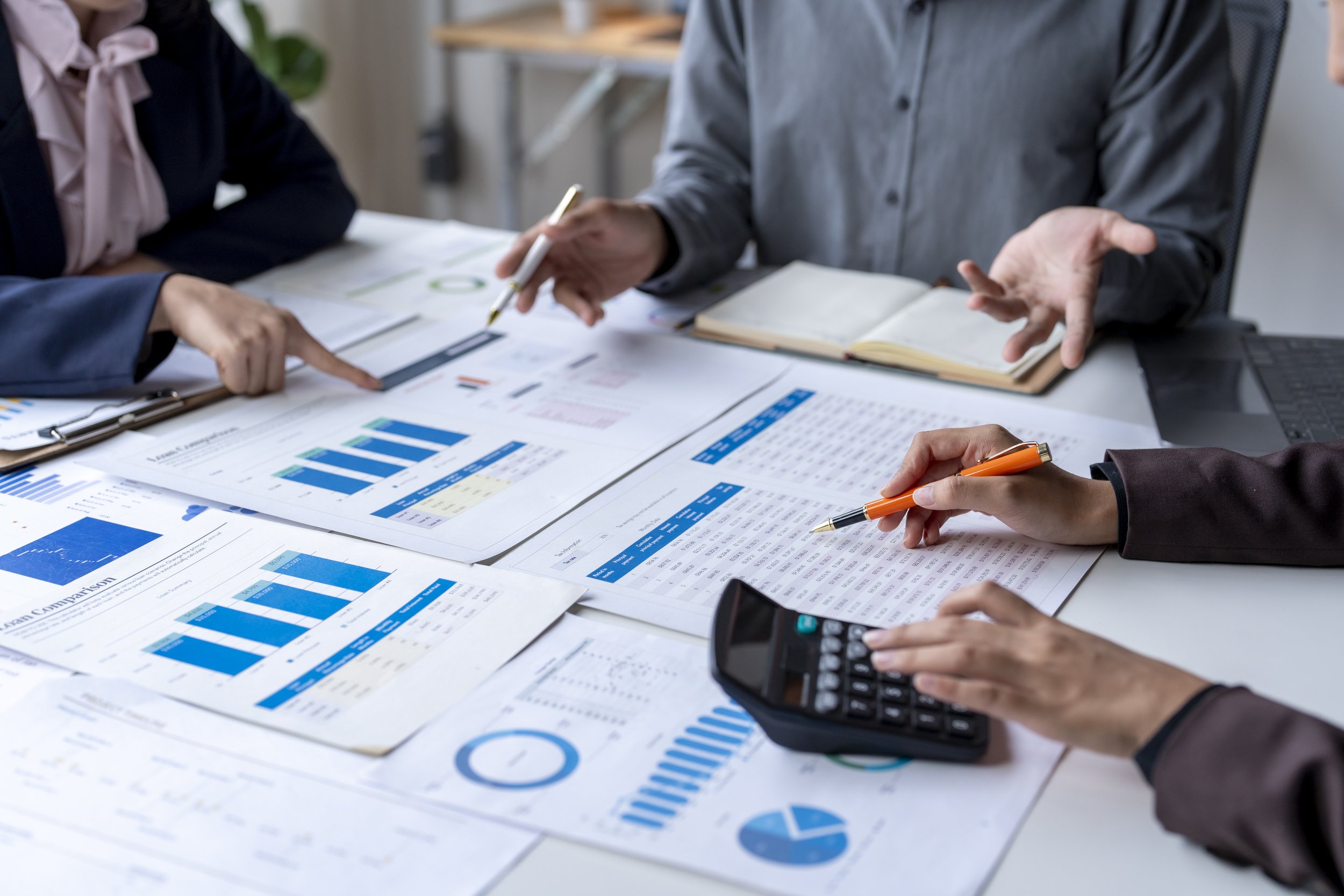 People in business attire discussing financial data, with printed charts, graphs, and spreadsheets on the table.