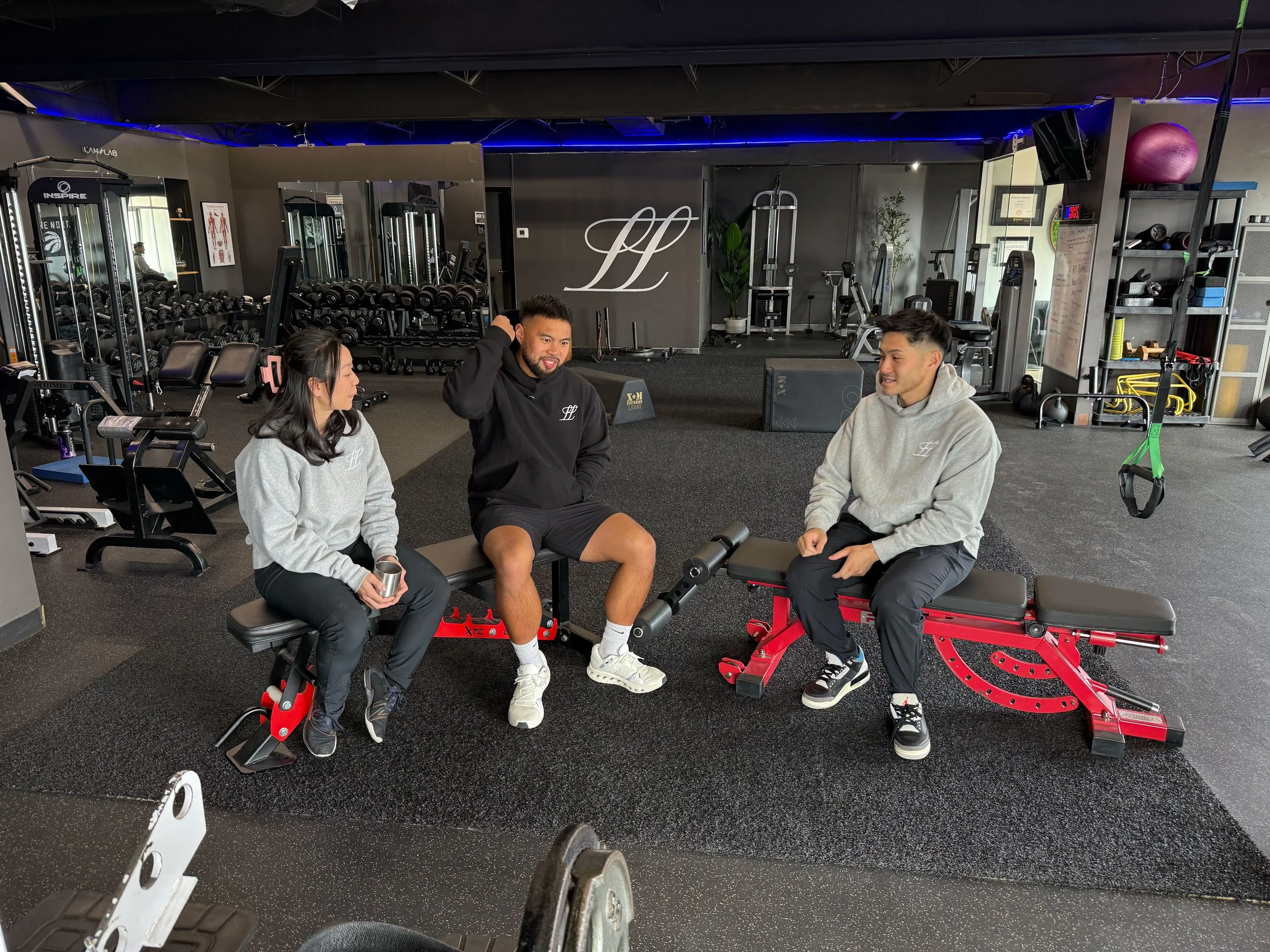 Commercial video production shoot inside a modern gym featuring three people filming branded fitness and lifestyle content surrounded by workout equipment.