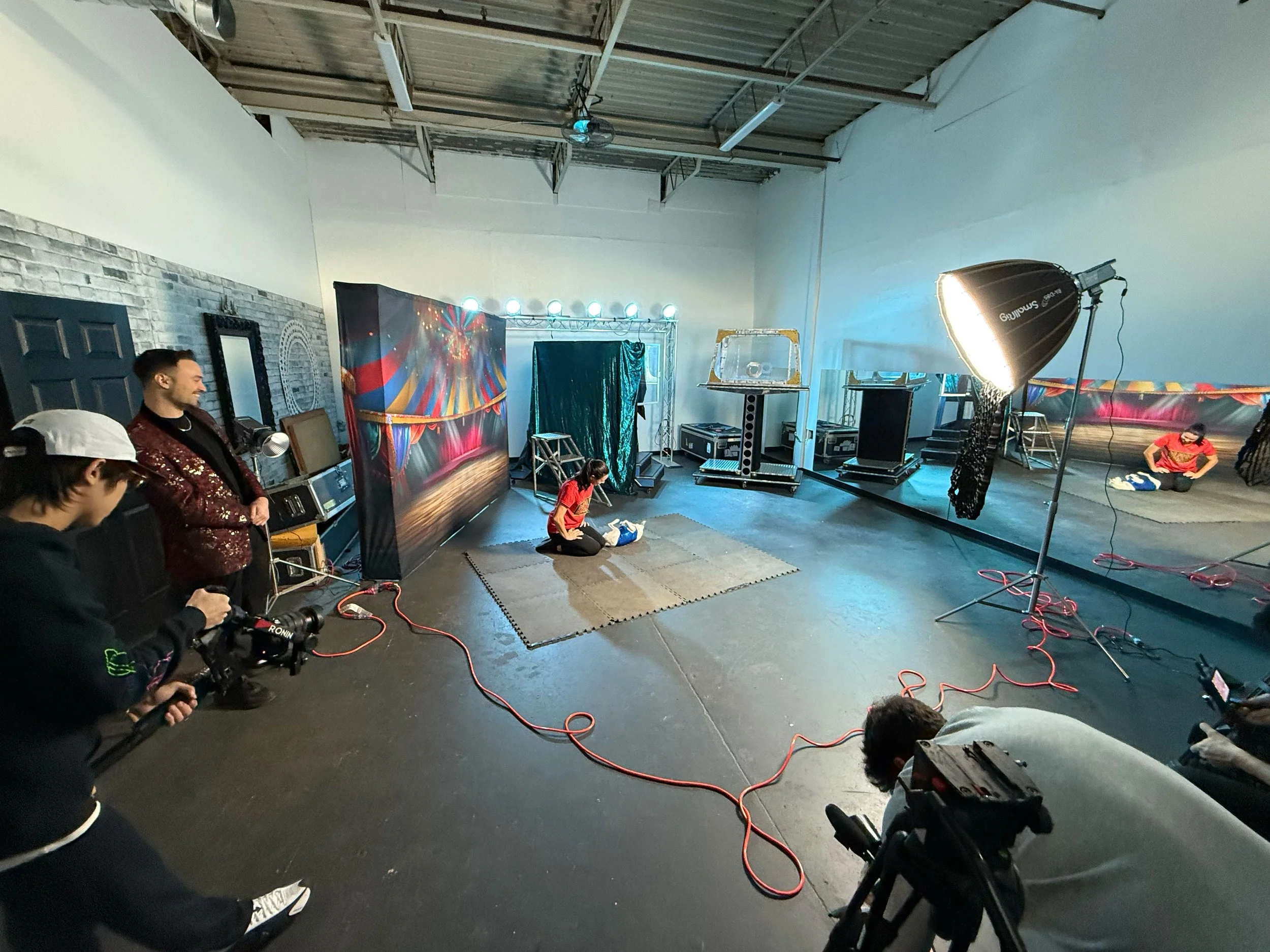Behind-the-scenes of a professional video shoot in a studio with camera operators, lighting equipment, colourful backdrops, and a performer rehearsing on set.