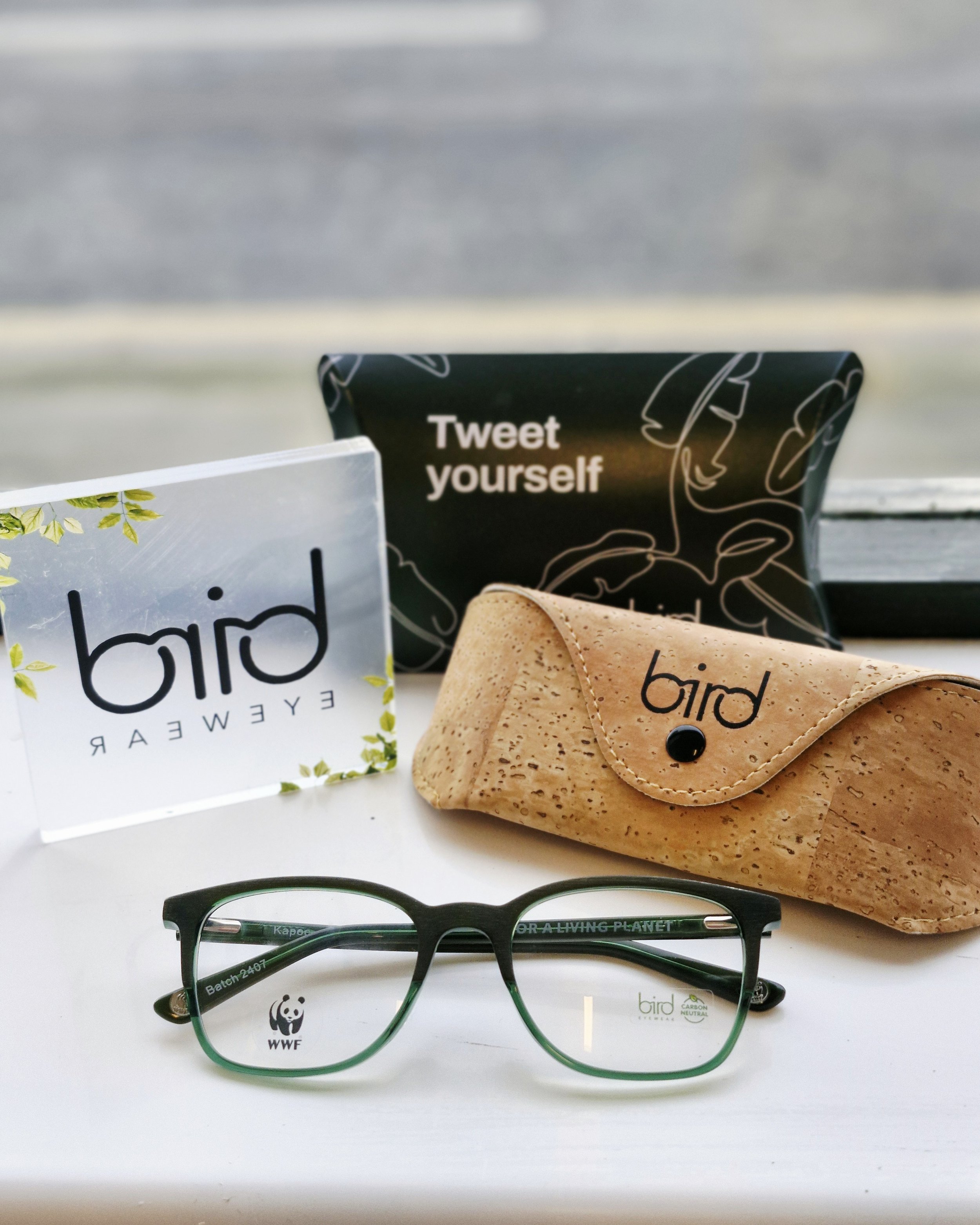 A pair of black and green eyeglasses, a wood-patterned eyeglasses case, a black pouch with white line art and the words 'Tweet yourself', and a clear card with black text and green leaf design reading 'bird eyewear' on a white surface.