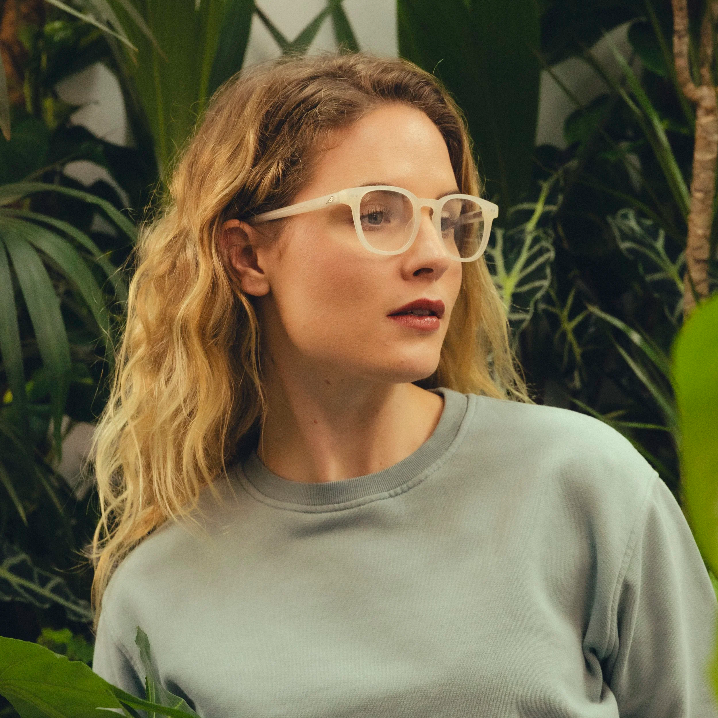 A young woman with curly blonde hair and glasses, looking to her left, surrounded by green foliage.