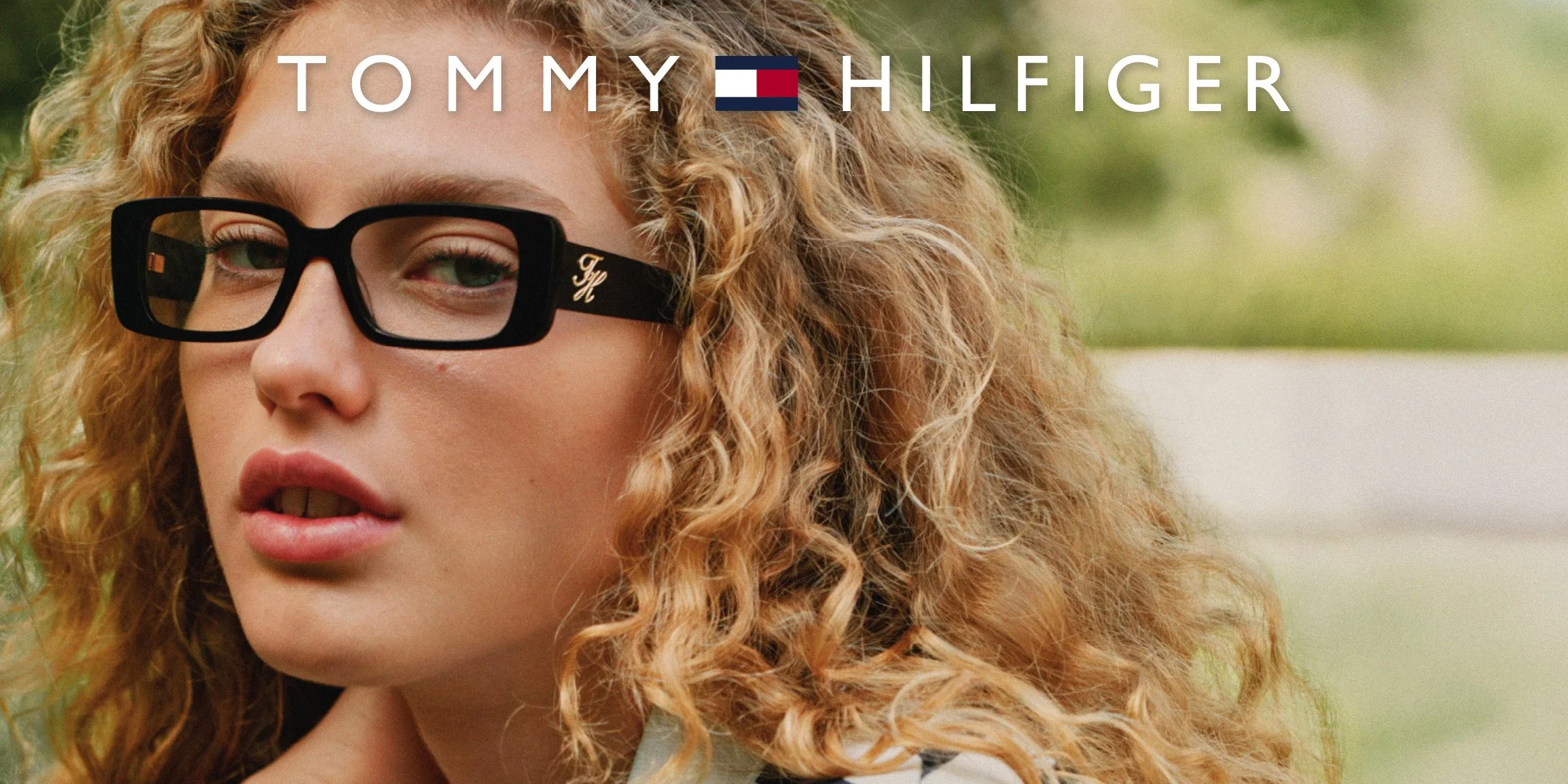 Close-up of a woman with curly blonde hair wearing black Tommy Hilfiger glasses, outdoors with a blurred background.